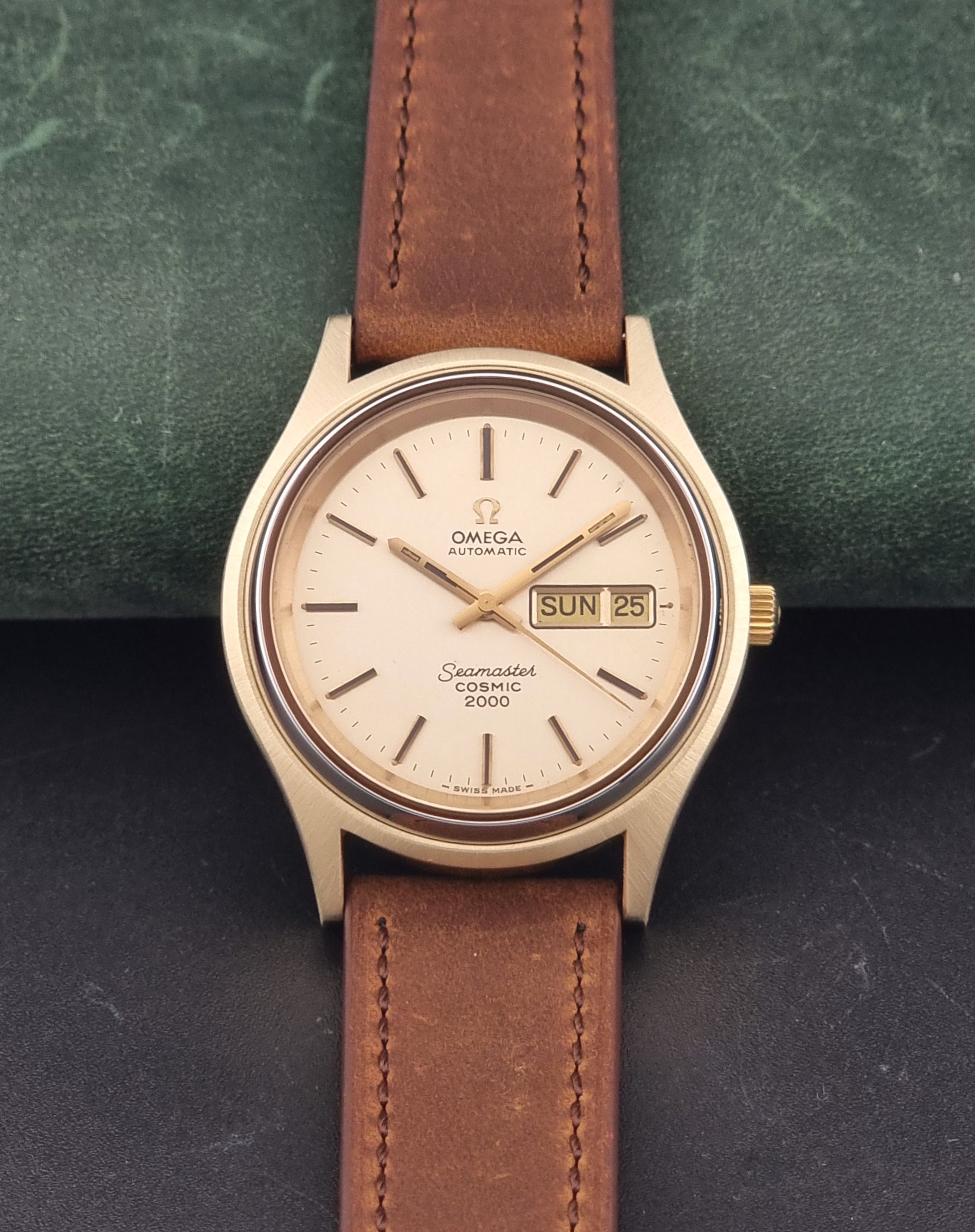 A Vintage Omega Wristwatch Seamaster Cosmic 2000 Jumbo 166.129 wristwatch with a gold-toned case, gold dial, and brown leather strap, displayed on a dark textured surface with a green background.
