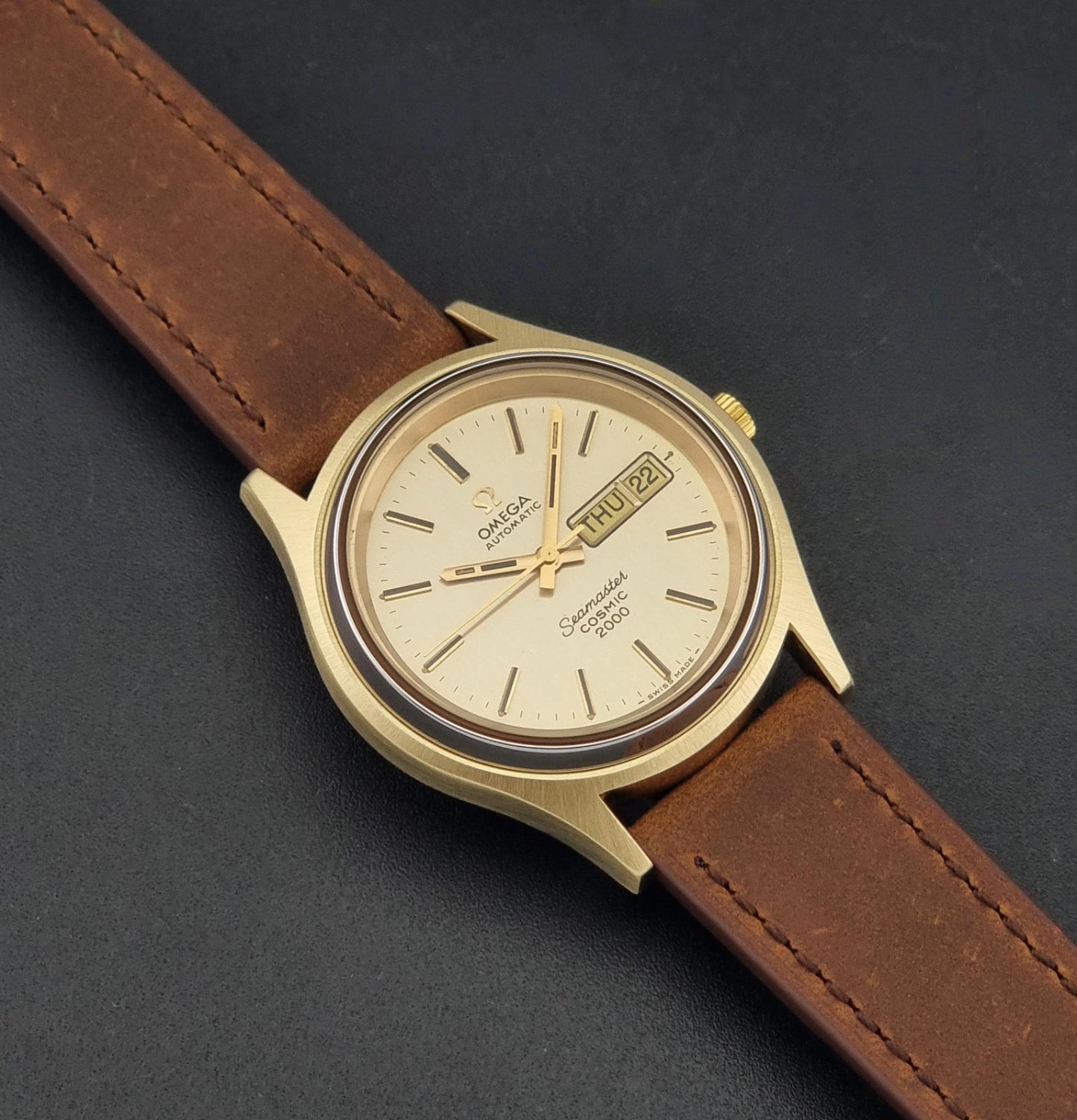 A Vintage Omega Wristwatch Seamaster Cosmic 2000 Jumbo 166.129 automatic watch with a gold-tone case, gold dial, and brown leather strap, photographed diagonally on a dark textured background.