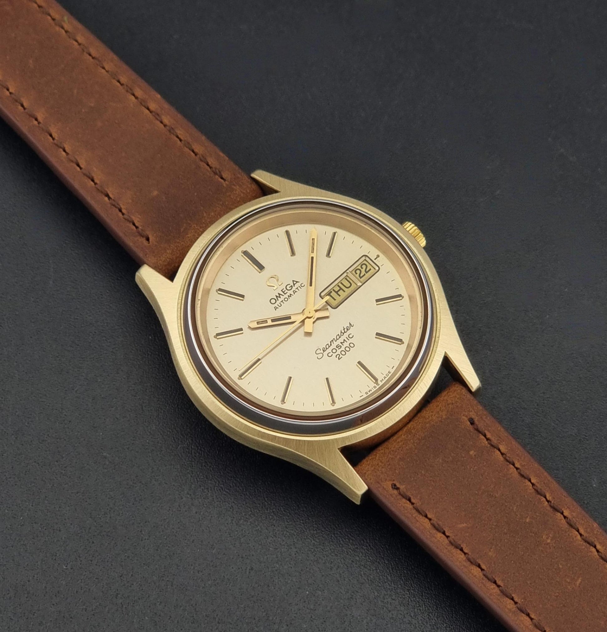 A Vintage Omega Wristwatch Seamaster Cosmic 2000 Jumbo 166.129 automatic watch with a gold-tone case, gold dial, and brown leather strap, photographed diagonally on a dark textured background.
