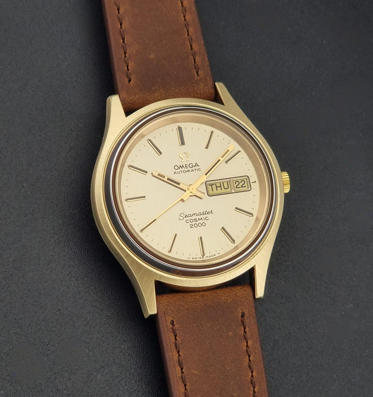 A Vintage Omega Wristwatch Seamaster Cosmic 2000 Jumbo 166.129 with a gold-tone case, gold dial, and brown leather strap, photographed on a dark textured background.