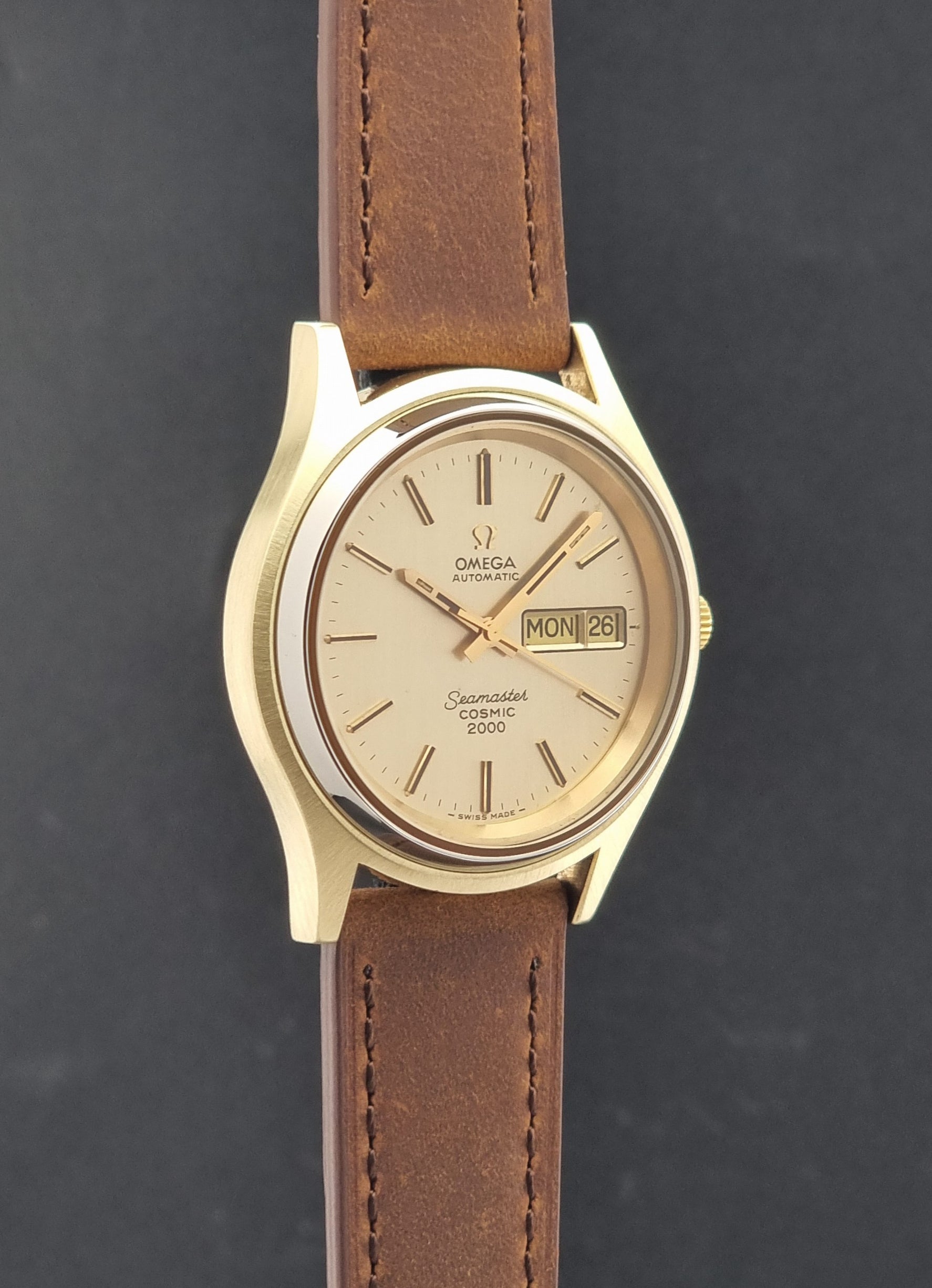 Vintage Omega Wristwatch Seamaster Cosmic 2000 Jumbo 166.129 with a gold-tone case, gold dial, and brown leather strap, shown against a dark gray background.