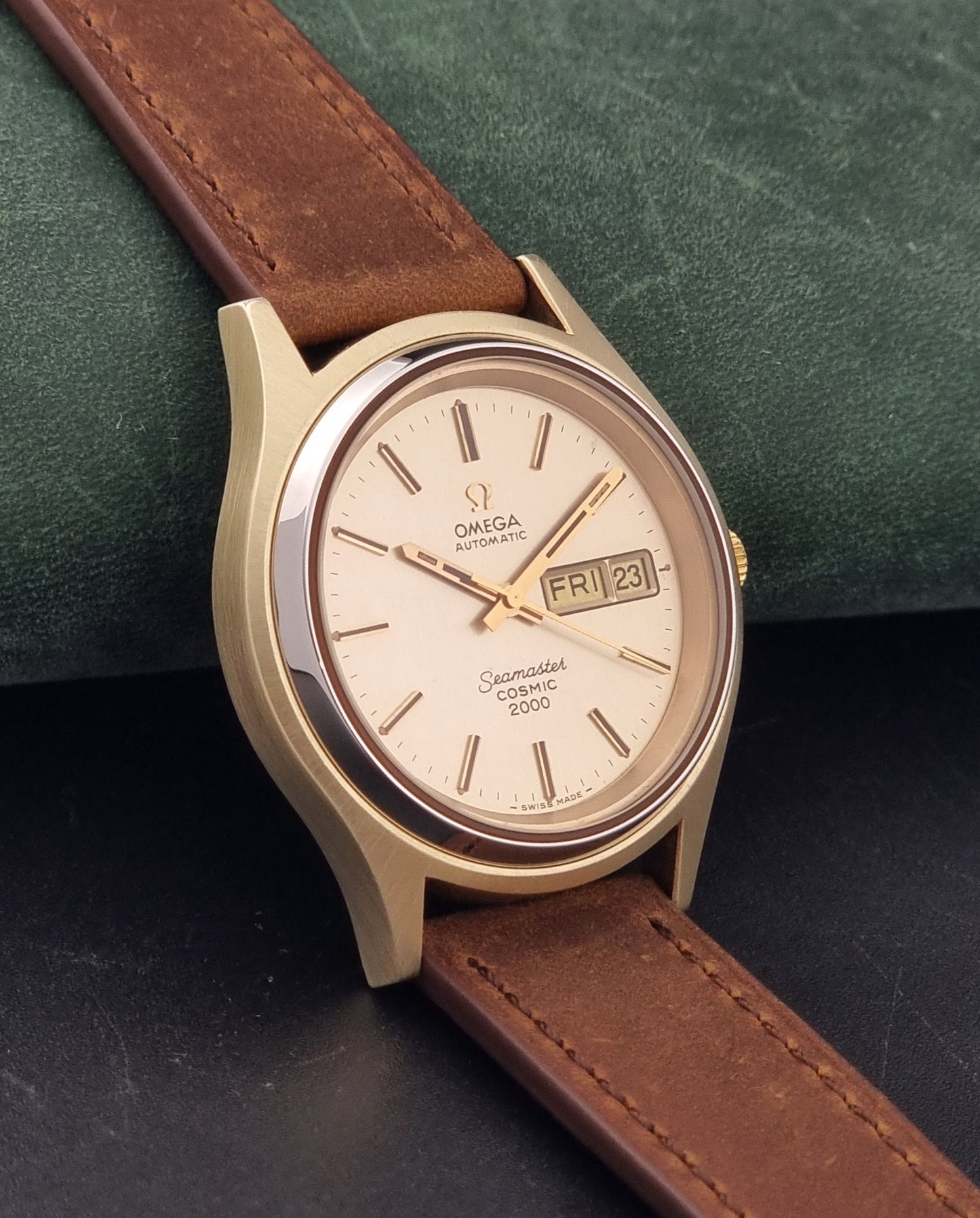 An Vintage Omega Wristwatch Seamaster Cosmic 2000 Jumbo 166.129 with a gold-tone case, gold dial, and brown leather strap, resting on a dark textured surface.