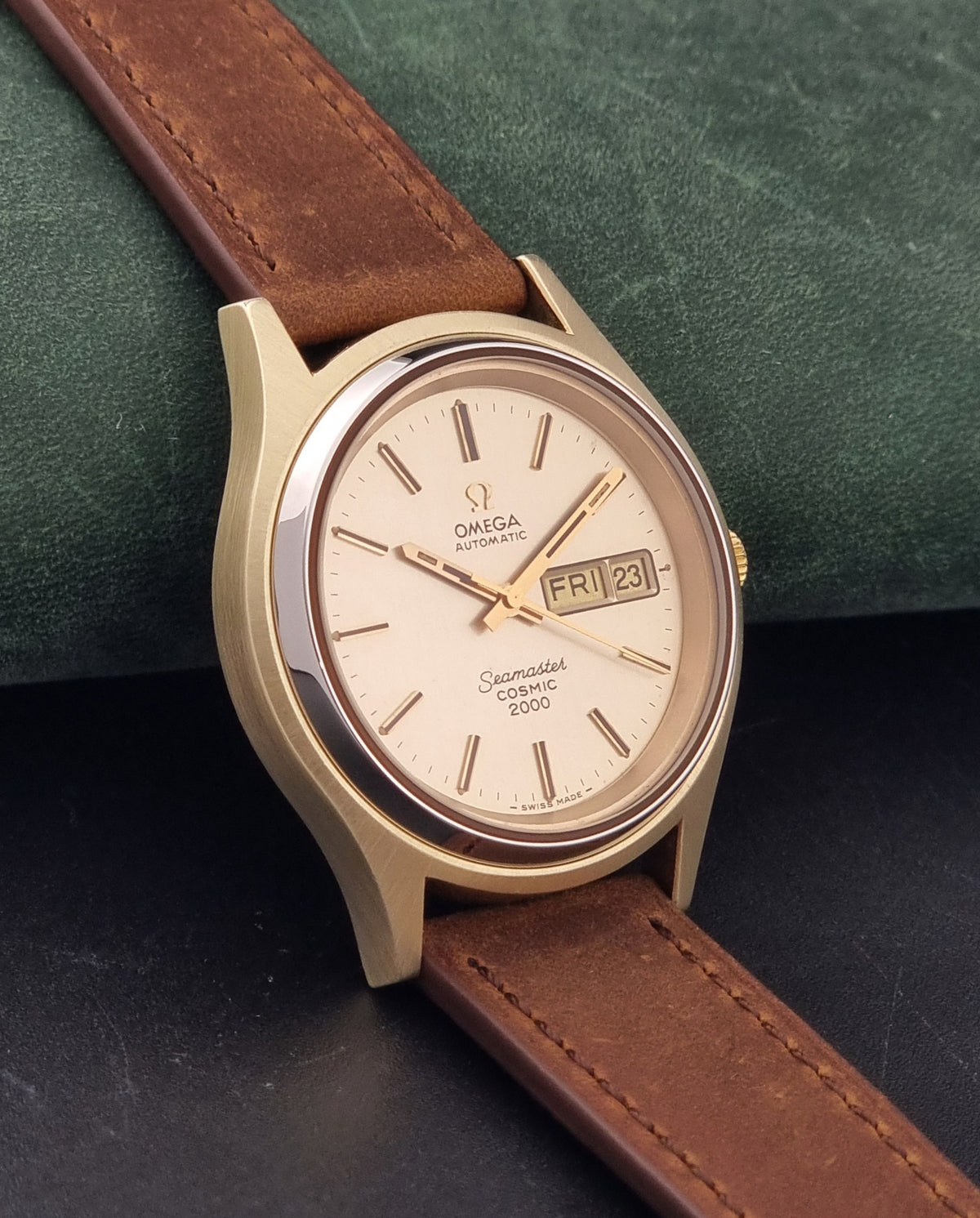 An Vintage Omega Wristwatch Seamaster Cosmic 2000 Jumbo 166.129 with a gold-tone case, gold dial, and brown leather strap, resting on a dark textured surface.