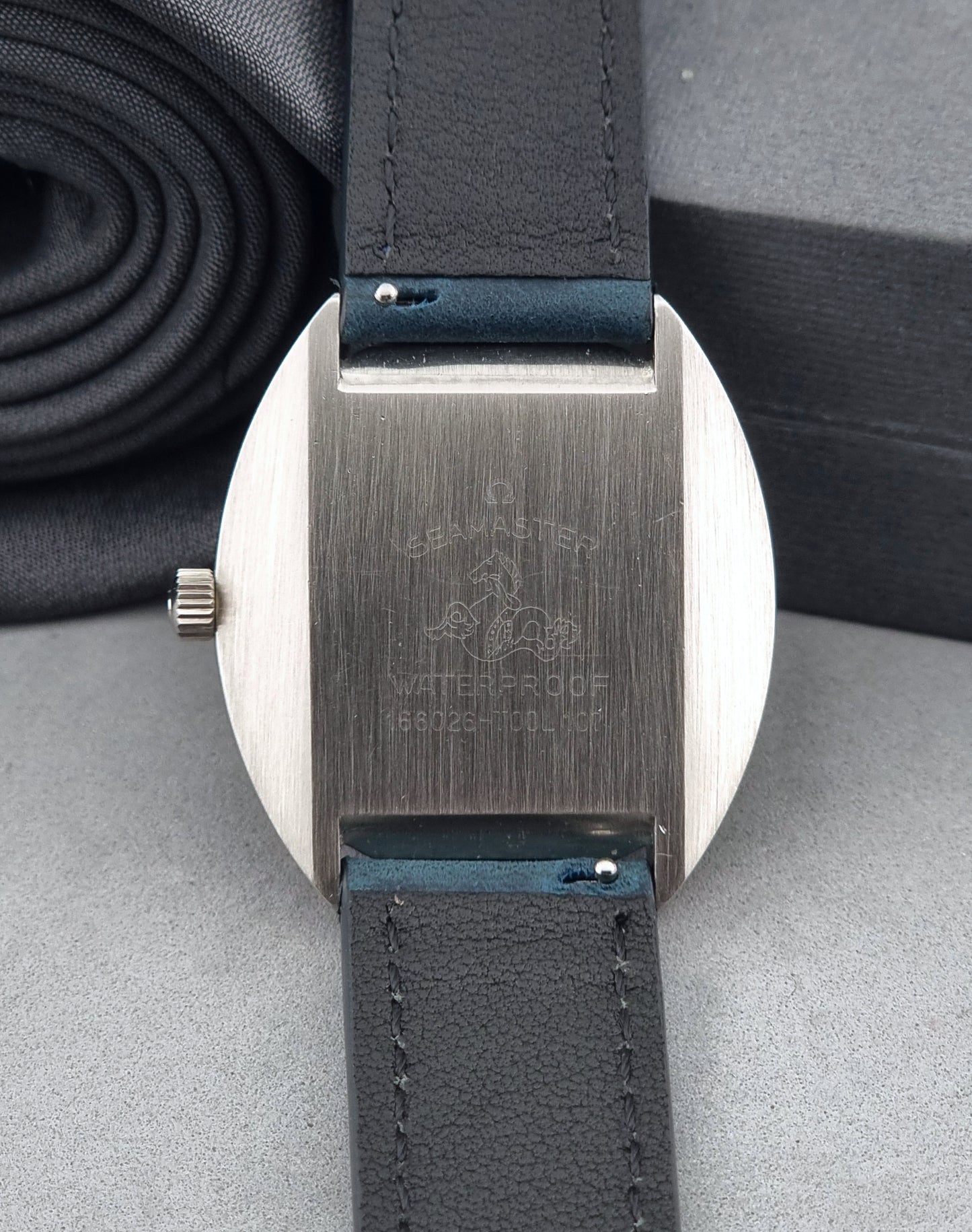 Vintage Omega Wristwatch 166.026 Seamaster Cosmic with blue leather strap on a dark surface with blue strap and silver case on a gray background case back view