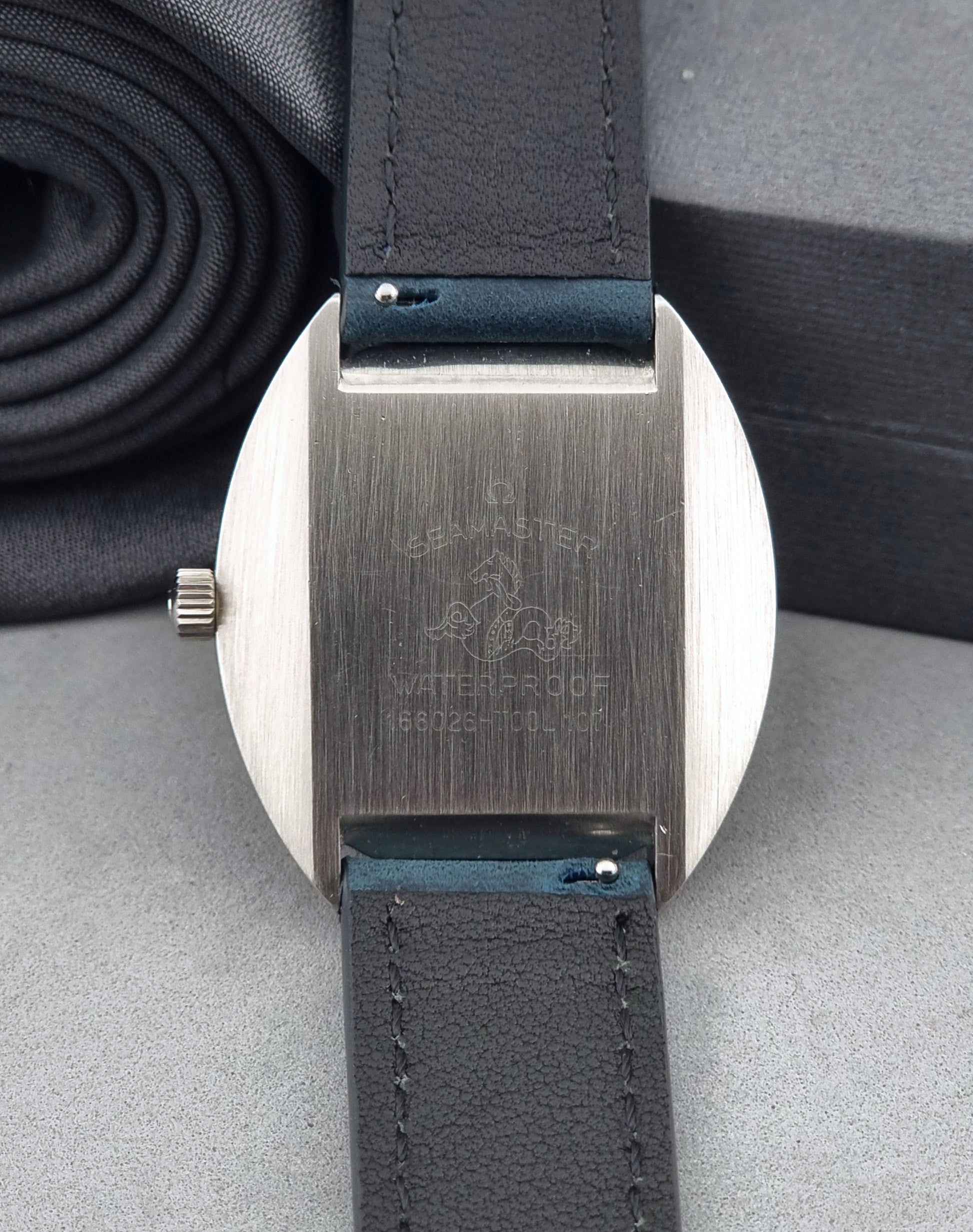 Vintage Omega Wristwatch 166.026 Seamaster Cosmic with blue leather strap on a dark surface with blue strap and silver case on a gray background case back view