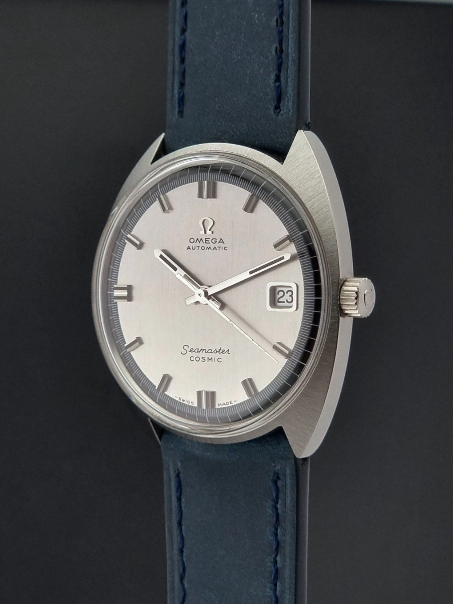 Vintage Omega Wristwatch 166.026 Seamaster Cosmic with blue leather strap on a dark surface with blue strap and silver case on a gray background close crown view