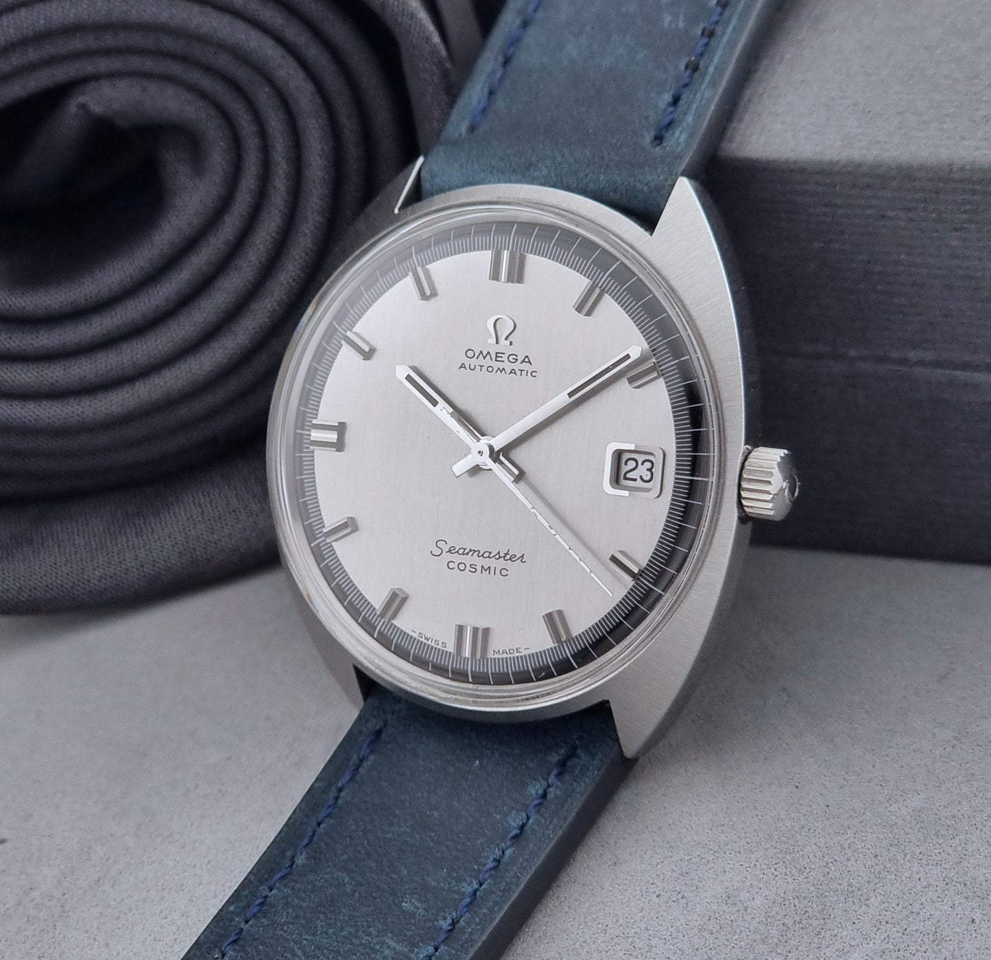 Vintage Omega Wristwatch 166.026 Seamaster Cosmic with blue leather strap on a dark surface with blue strap and silver case on a gray background crown view