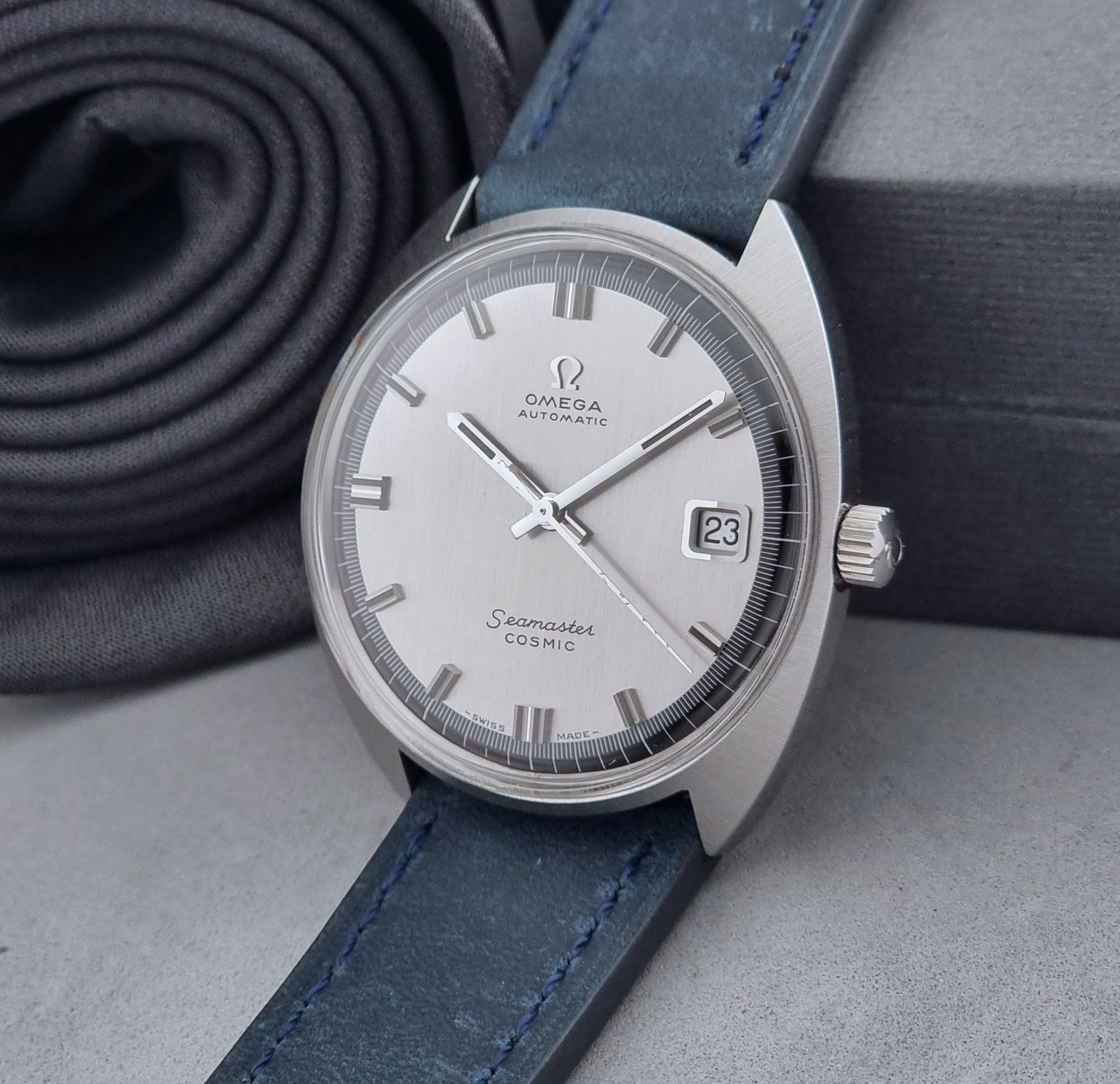Vintage Omega Wristwatch 166.026 Seamaster Cosmic with blue leather strap on a dark surface with blue strap and silver case on a gray background crown view
