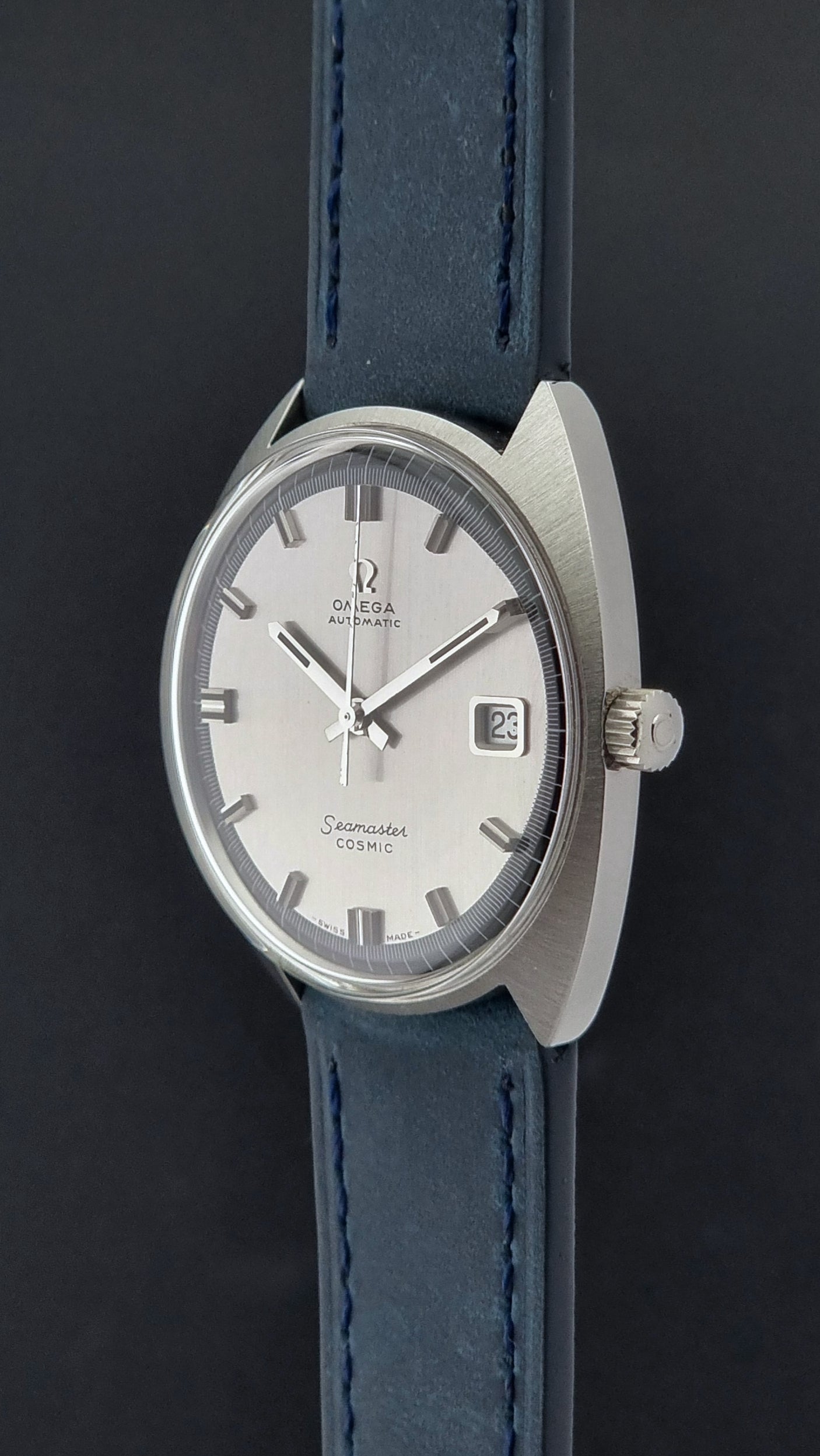 Vintage Omega Wristwatch 166.026 Seamaster Cosmic with blue leather strap on a dark surface with blue strap and silver case on a gray background crown profile view