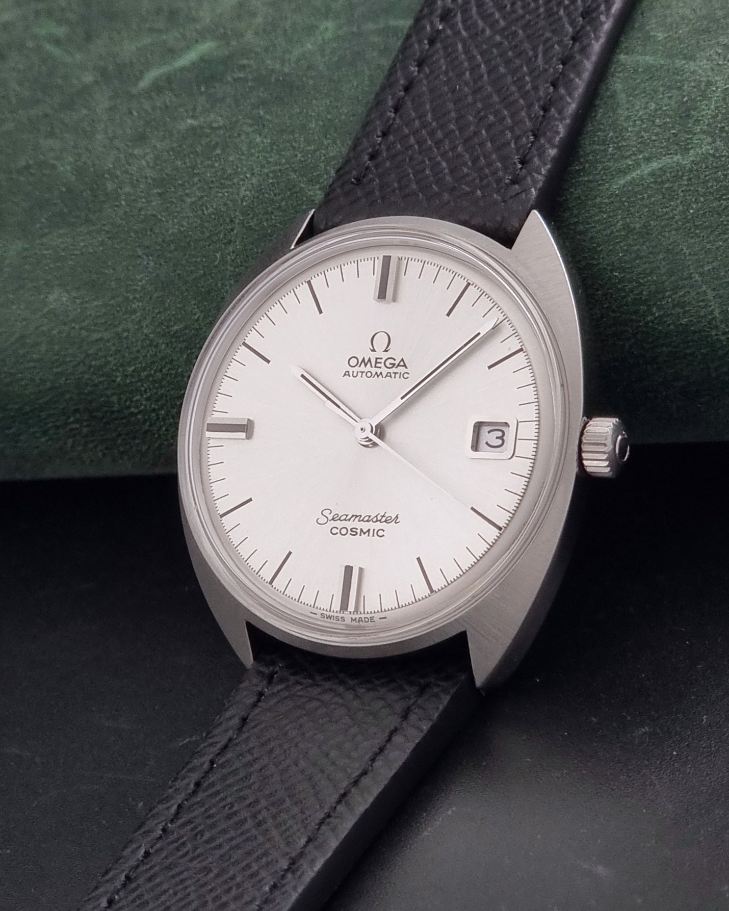 Vintage Omega 166.026 Seamaster Cosmic Automatic watch with a silver-tone case, silver dial, and textured black leather strap, on a dark background crown view
