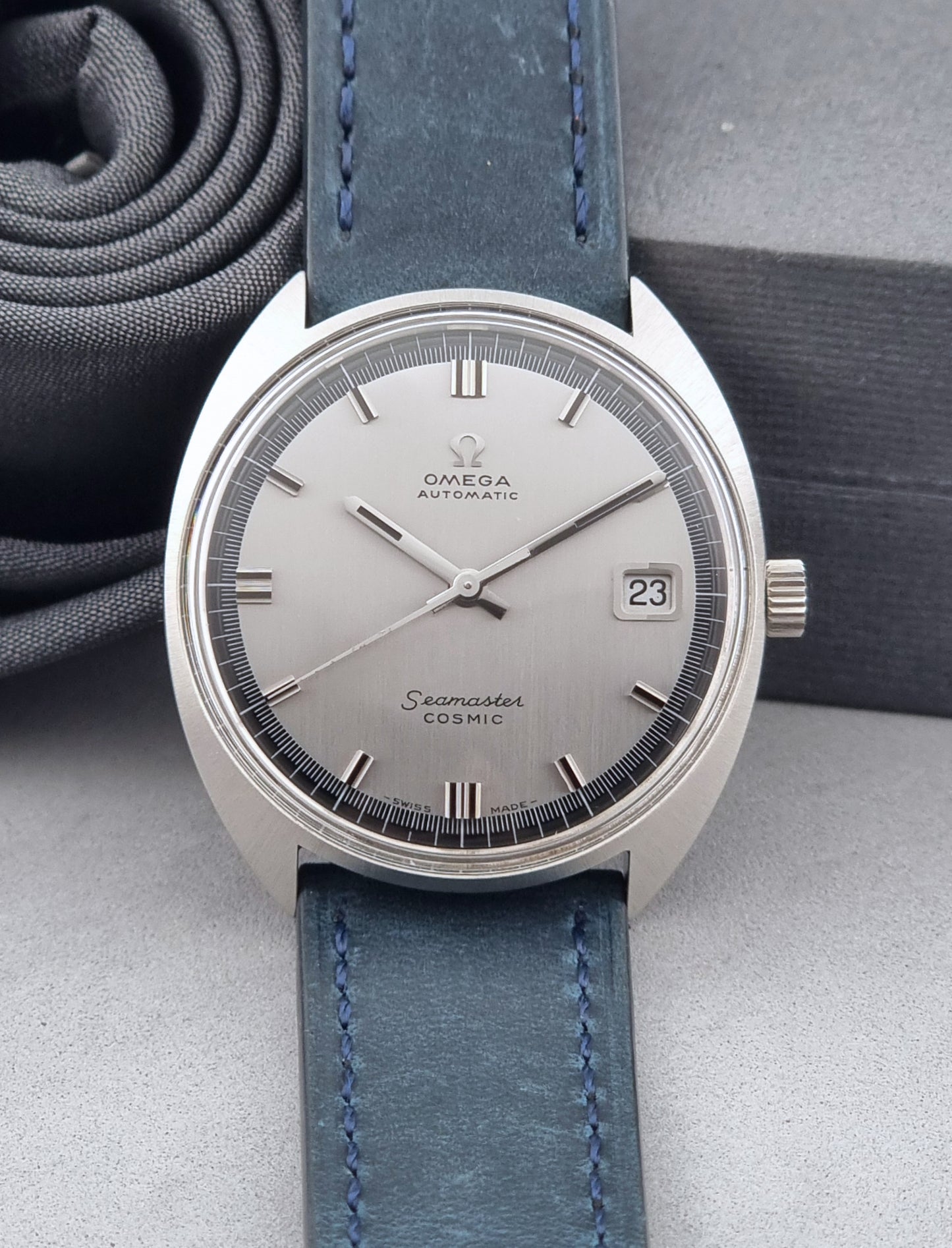 Vintage Omega Wristwatch 166.026 Seamaster Cosmic with blue leather strap on a dark surface with blue strap and silver case on a gray background front view