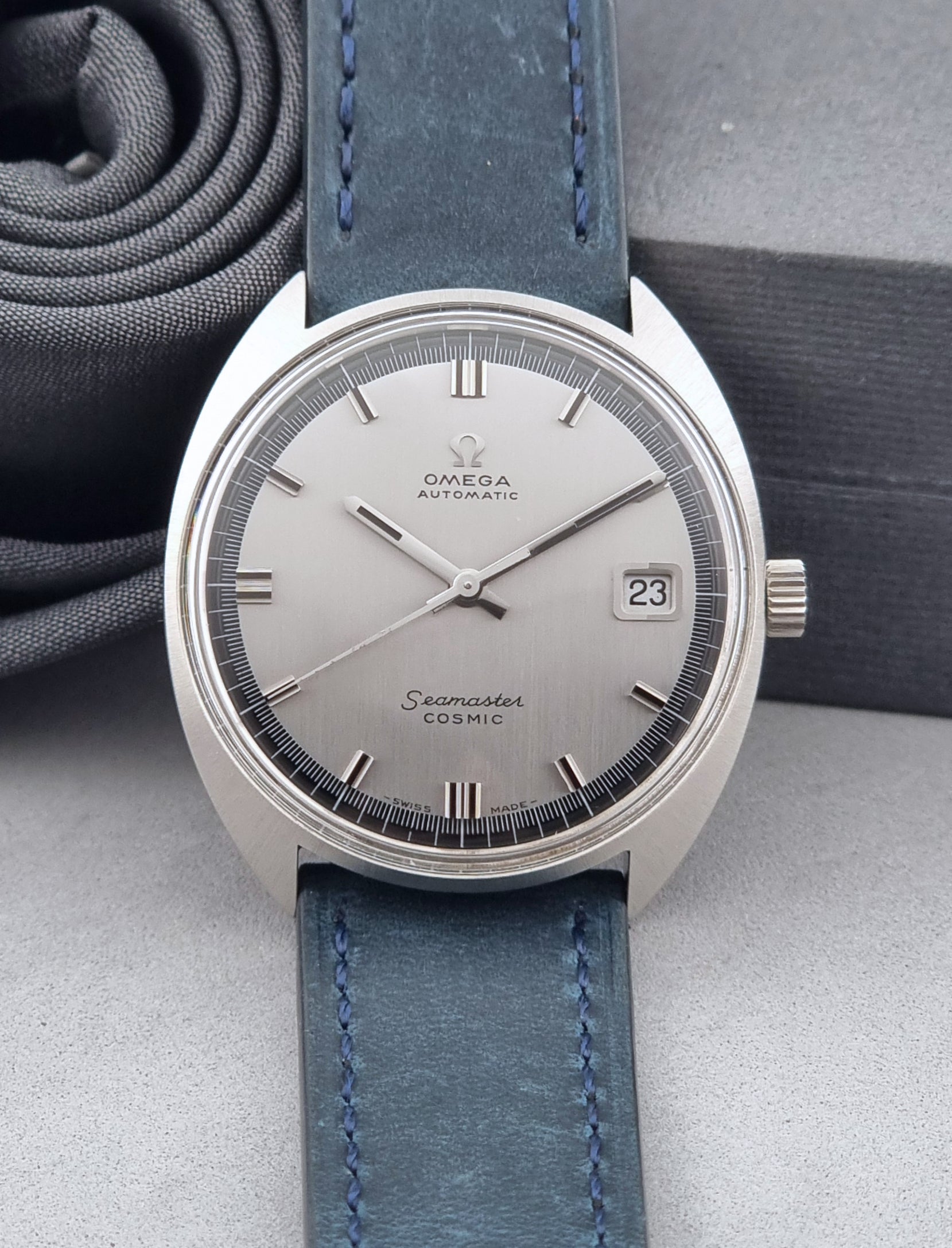 Vintage Omega Wristwatch 166.026 Seamaster Cosmic with blue leather strap on a dark surface with blue strap and silver case on a gray background front view