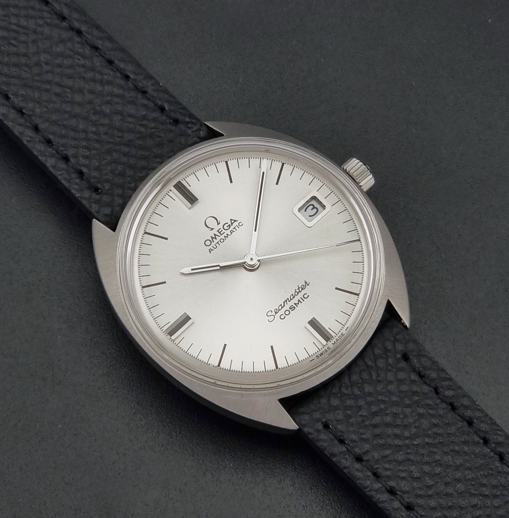 Vintage Omega 166.026 Seamaster Cosmic Automatic watch with a silver-tone case, silver dial, and textured black leather strap, on a dark background close up dial view