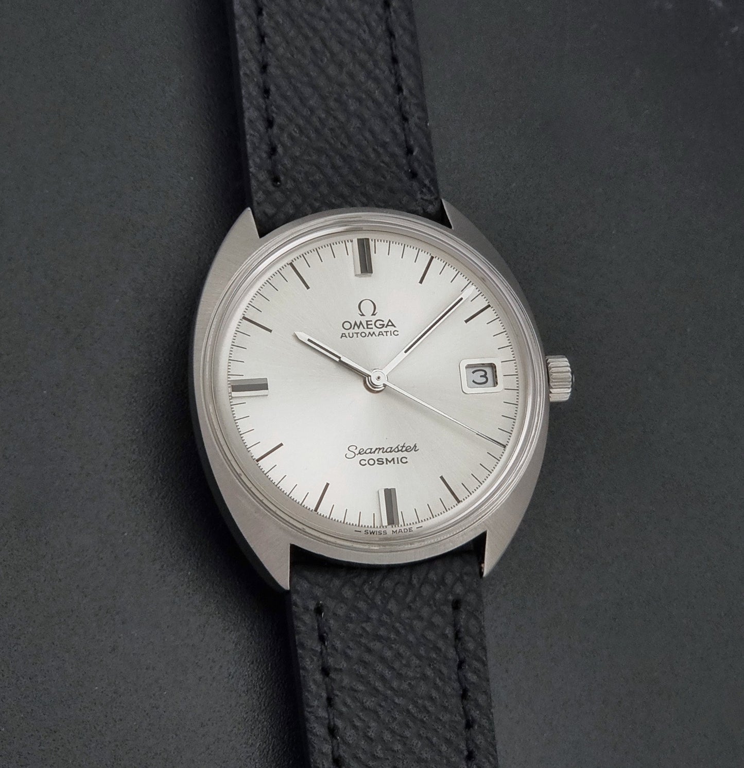 Vintage Omega 166.026 Seamaster Cosmic Automatic watch with a silver-tone case, silver dial, and textured black leather strap, on a dark background front view