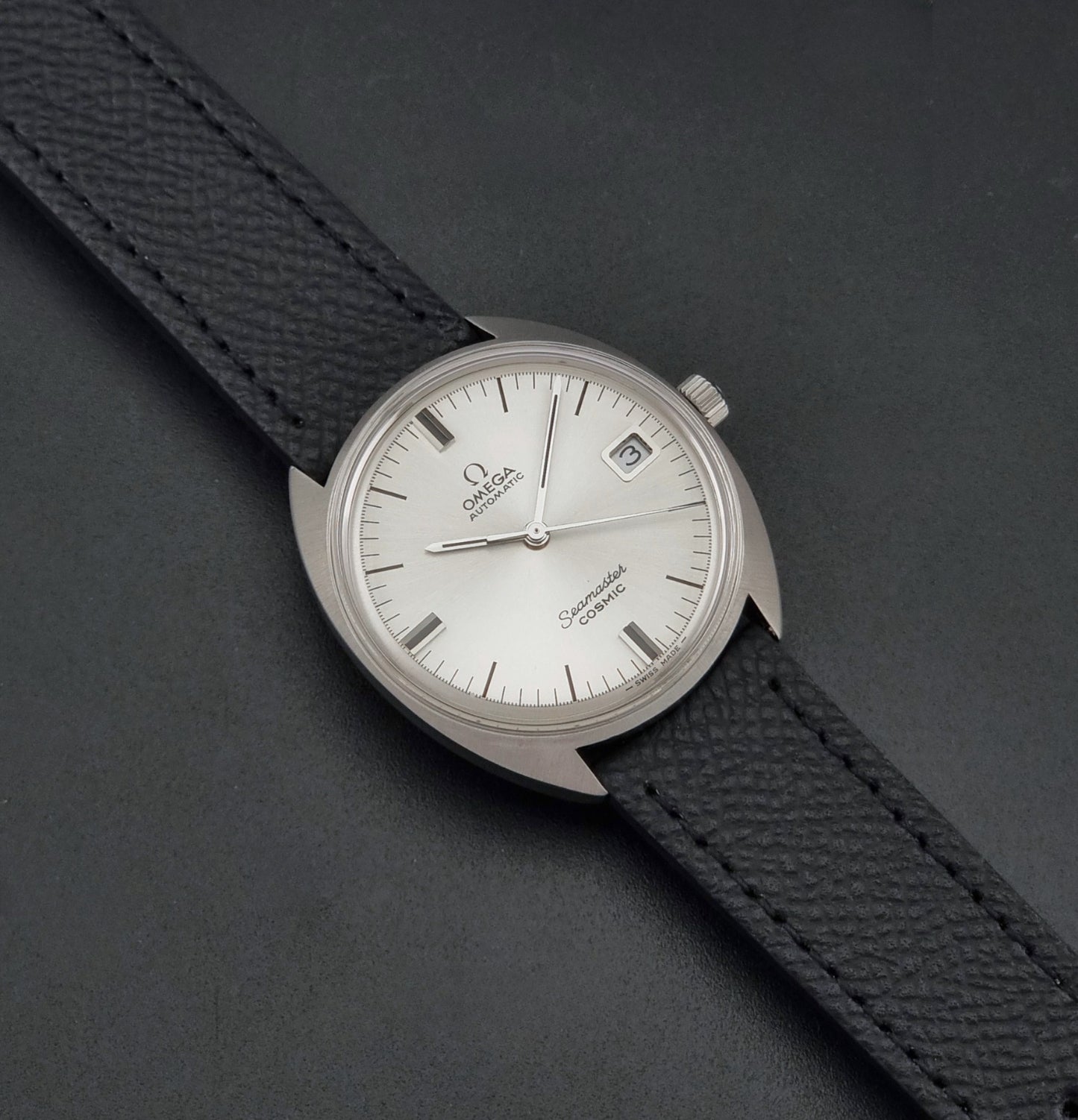 Vintage Omega 166.026 Seamaster Cosmic Automatic watch with a silver-tone case, silver dial, and textured black leather strap, on a dark background dial