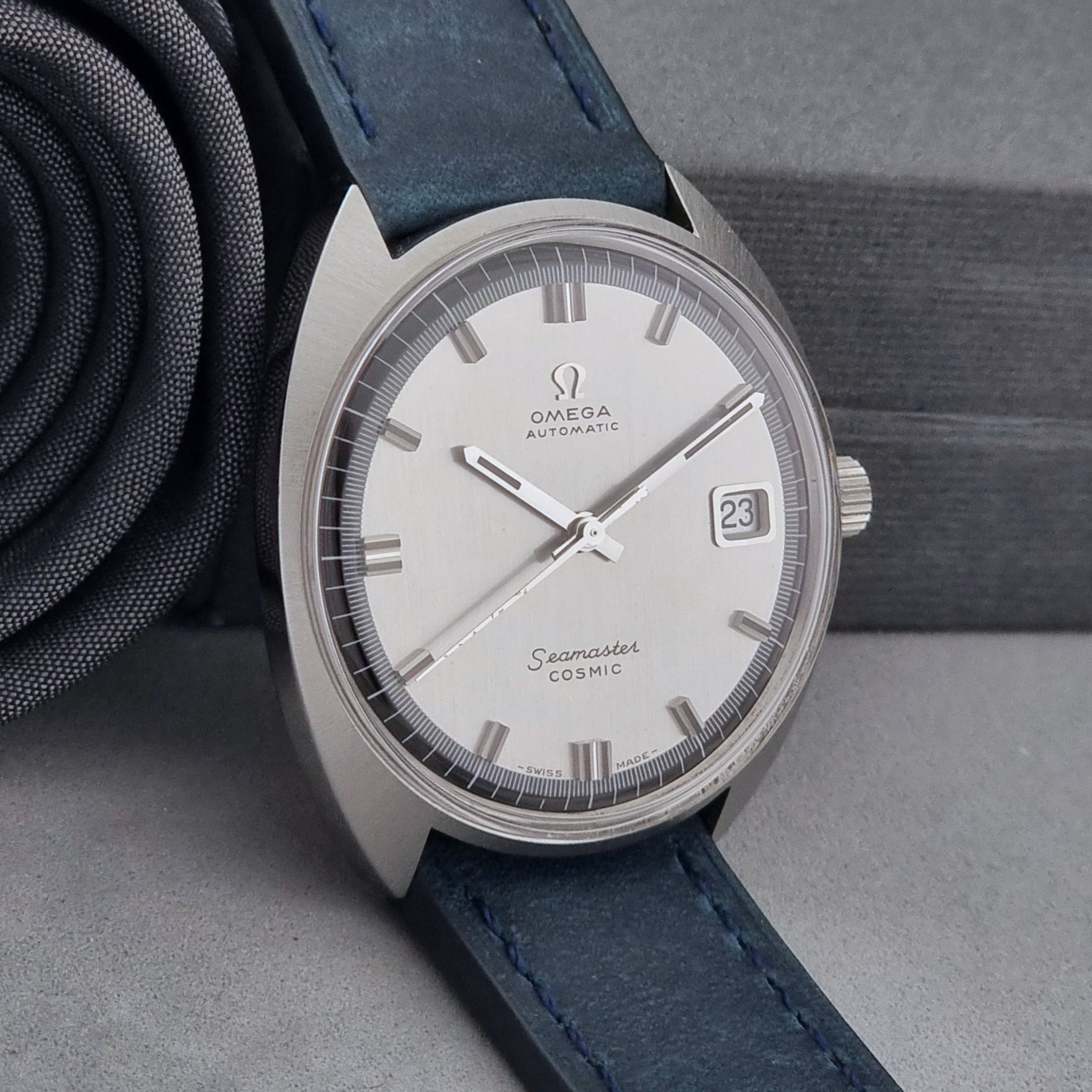 Vintage Omega Wristwatch 166.026 Seamaster Cosmic with blue leather strap on a dark surface with blue strap and silver case on a gray background side view