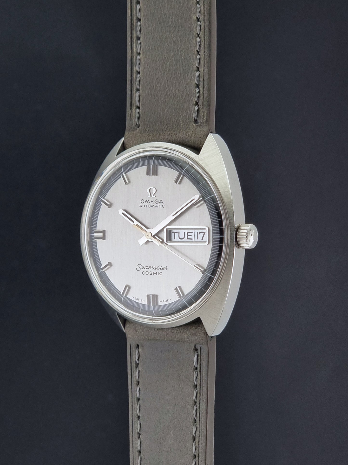 Vintage Omega 166.036 Seamaster Cosmic watch with a silver-toned case, a light gray dial with a day-date window, and a gray leather strap, on a dark background crown profile view