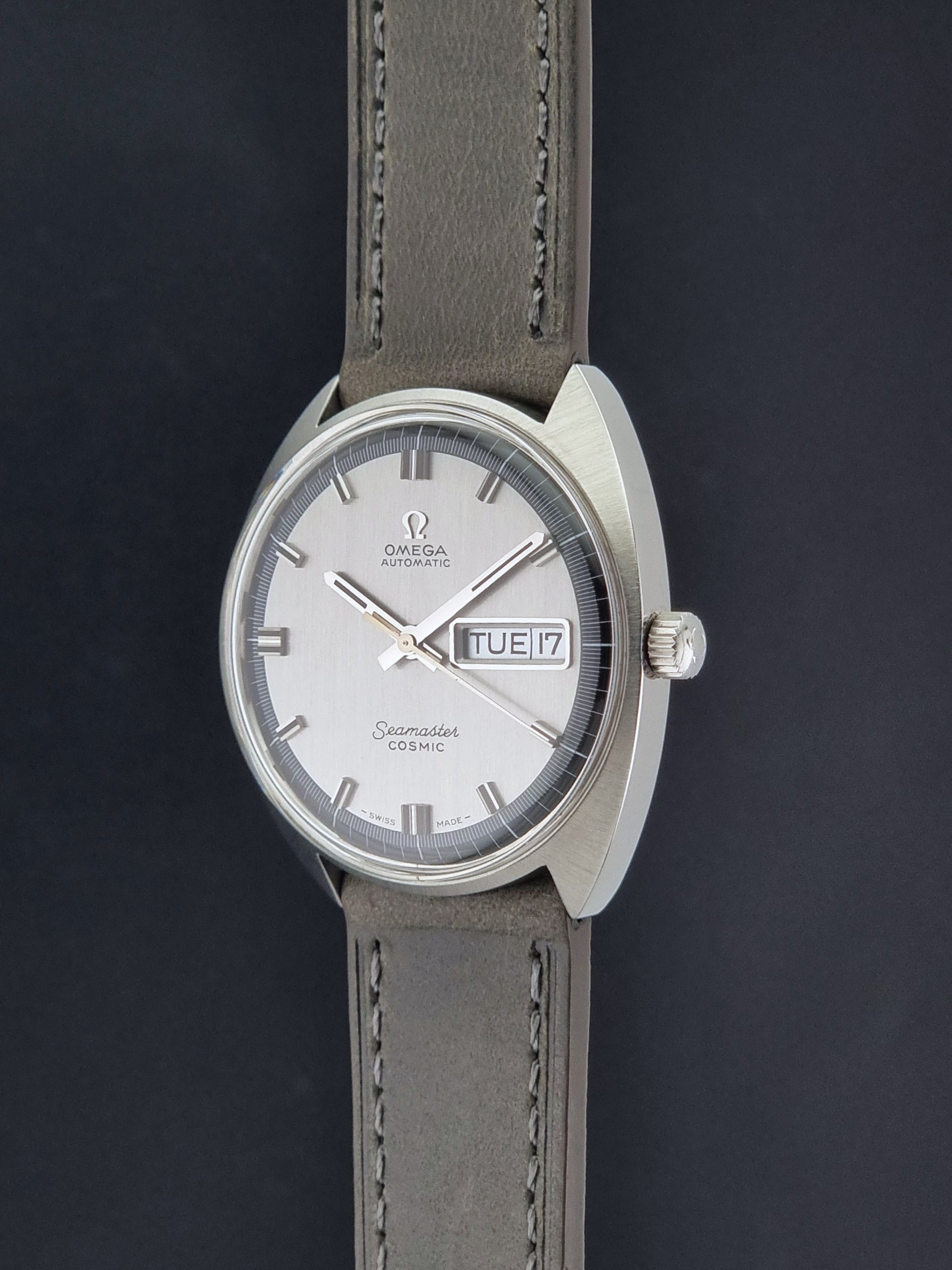 Vintage Omega 166.036 Seamaster Cosmic watch with a silver-toned case, a light gray dial with a day-date window, and a gray leather strap, on a dark background crown profile view