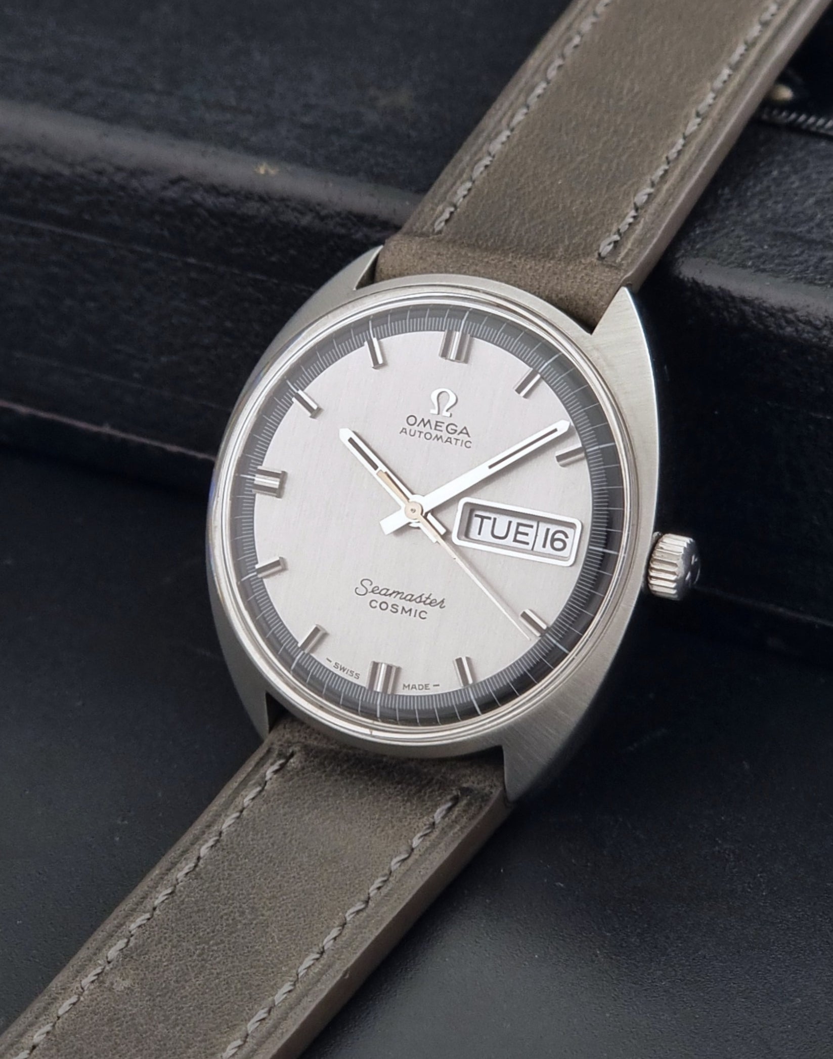 Vintage Omega 166.036 Seamaster Cosmic watch with a silver-toned case, a light gray dial with a day-date window, and a gray leather strap, on a dark background crown view