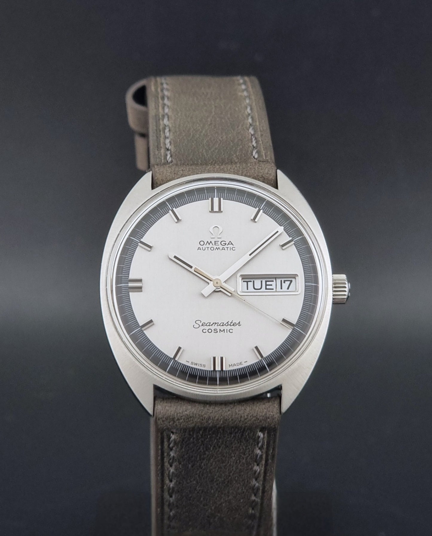Vintage Omega 166.036 Seamaster Cosmic watch with a silver-toned case, a light gray dial with a day-date window, and a gray leather strap, on a dark background close dial view