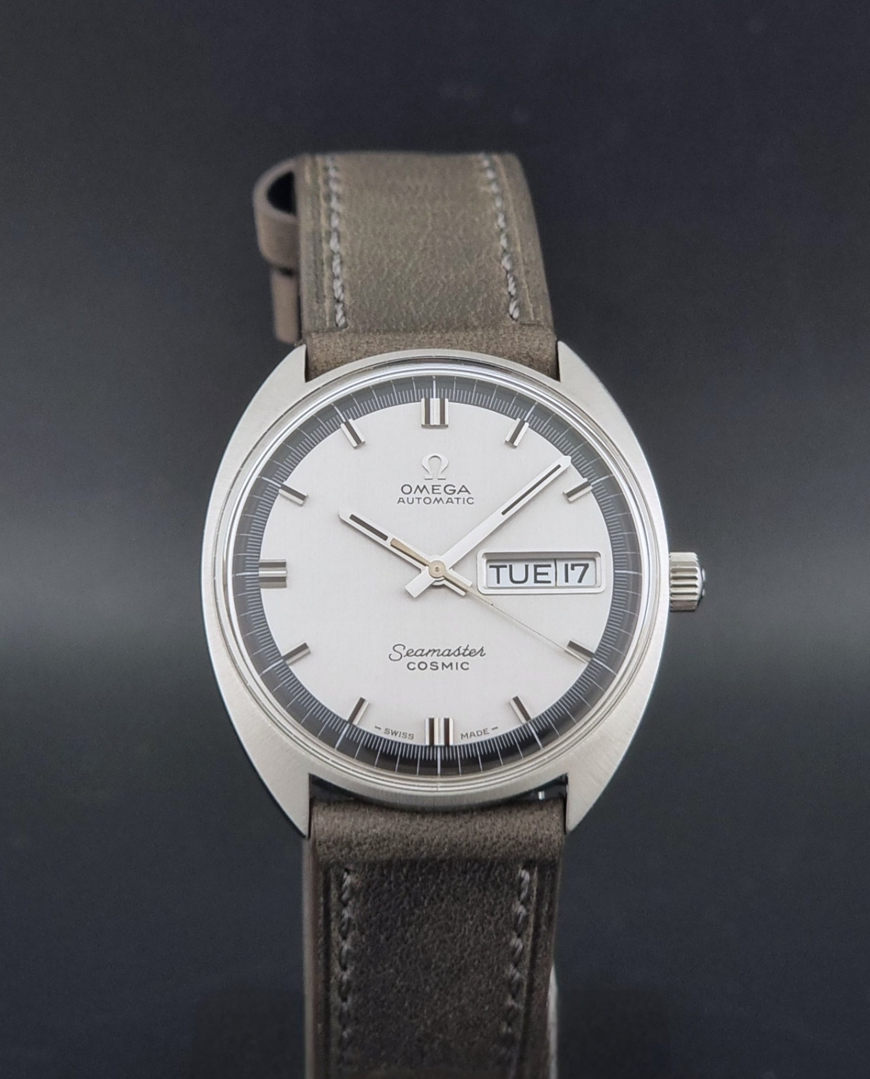 Vintage Omega 166.036 Seamaster Cosmic watch with a silver-toned case, a light gray dial with a day-date window, and a gray leather strap, on a dark background close dial view