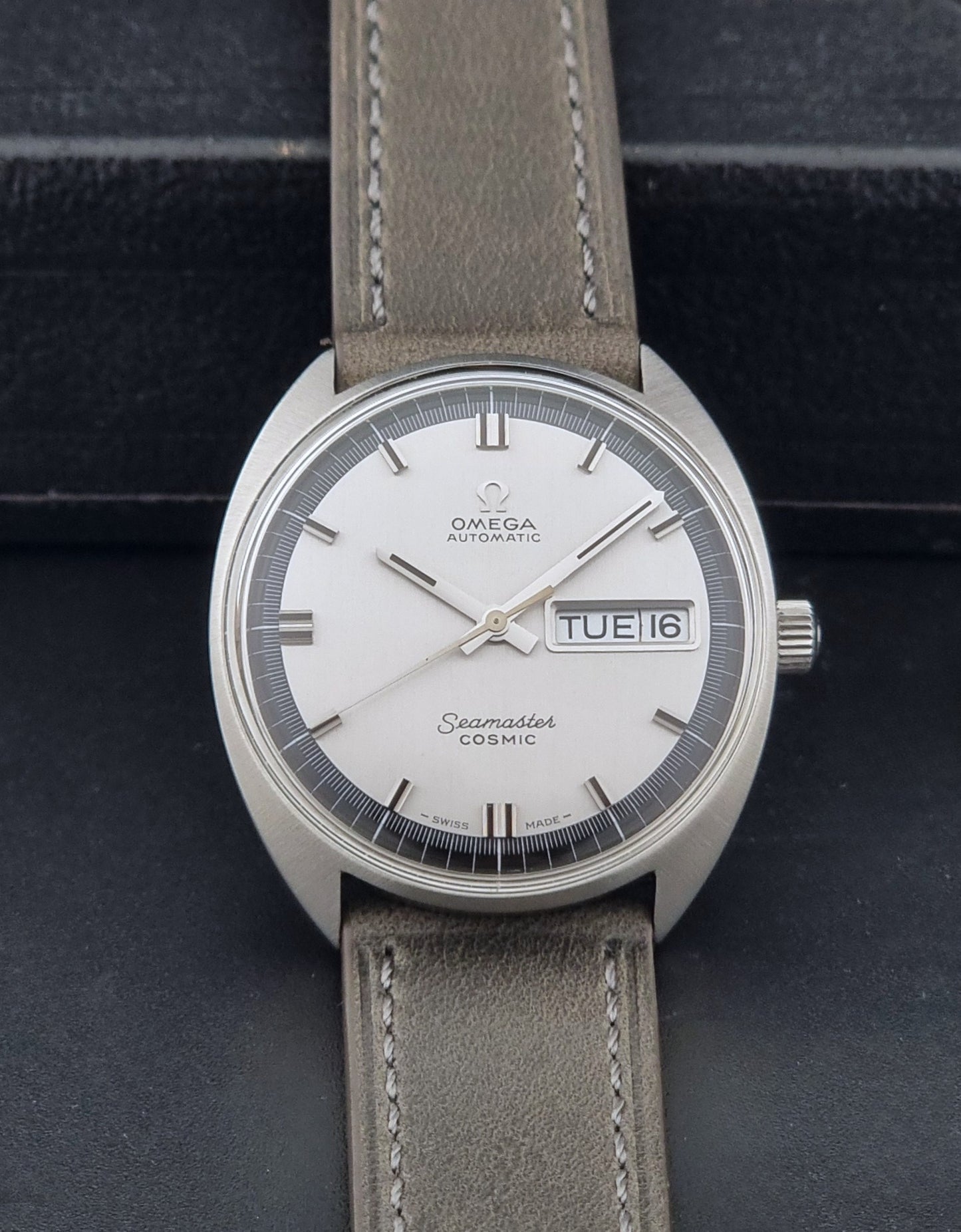 Vintage Omega 166.036 Seamaster Cosmic watch with a silver-toned case, a light gray dial with a day-date window, and a gray leather strap, on a dark background dial view