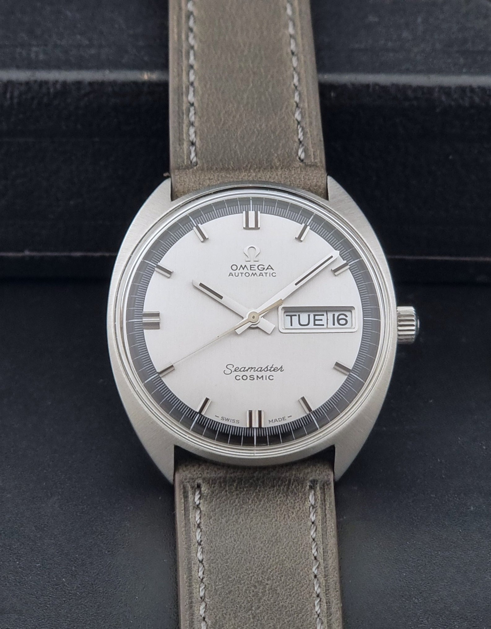 Vintage Omega 166.036 Seamaster Cosmic watch with a silver-toned case, a light gray dial with a day-date window, and a gray leather strap, on a dark background dial view