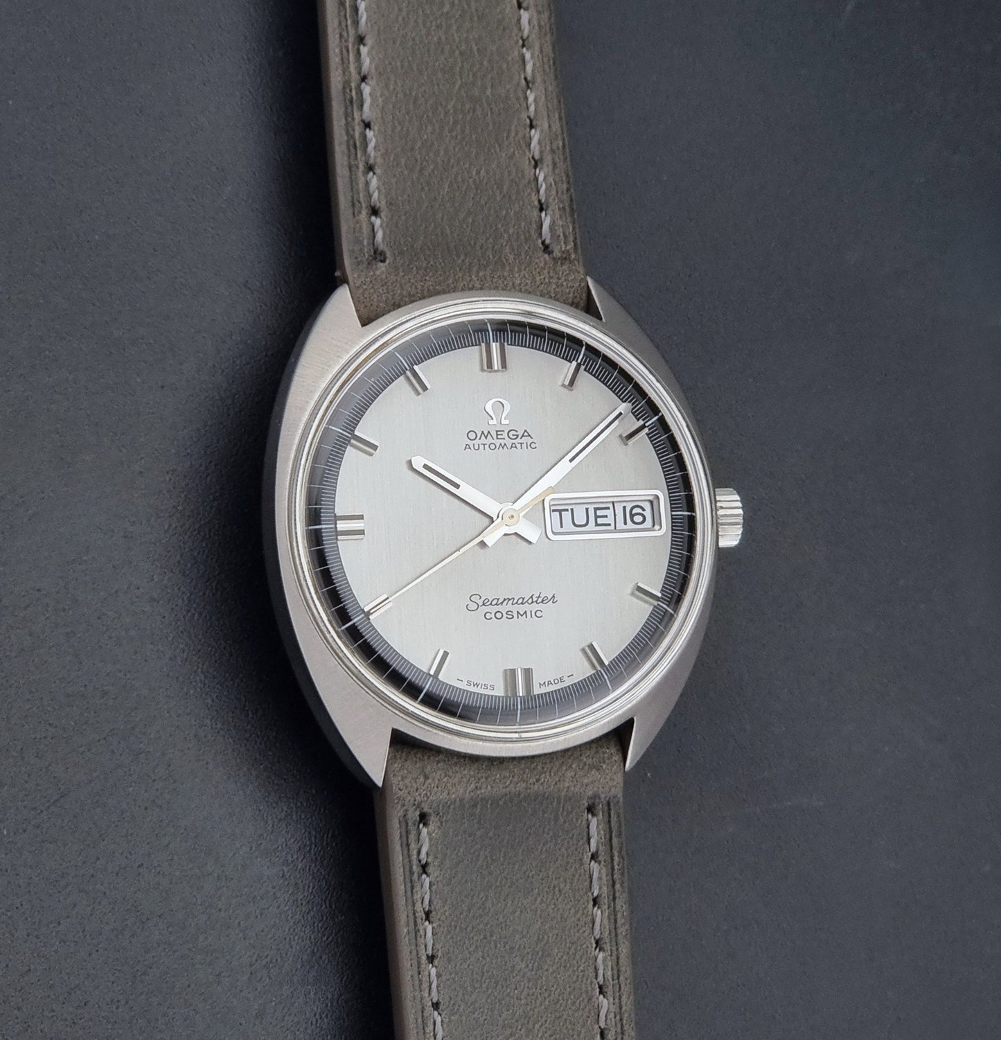 Vintage Omega 166.036 Seamaster Cosmic watch with a silver-toned case, a light gray dial with a day-date window, and a gray leather strap, on a dark background front view