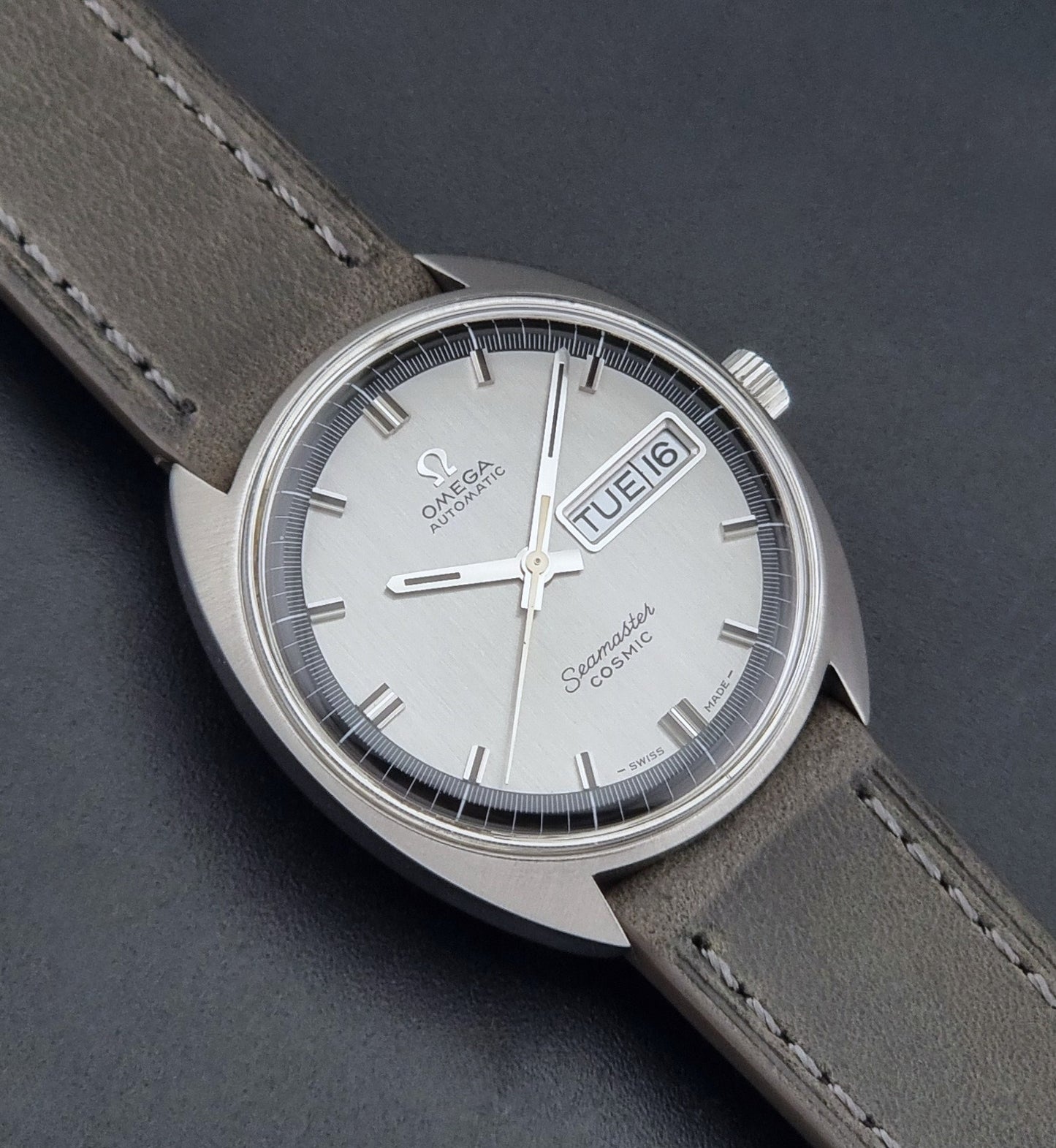 Vintage Omega 166.036 Seamaster Cosmic watch with a silver-toned case, a light gray dial with a day-date window, and a gray leather strap, on a dark background close up dial view