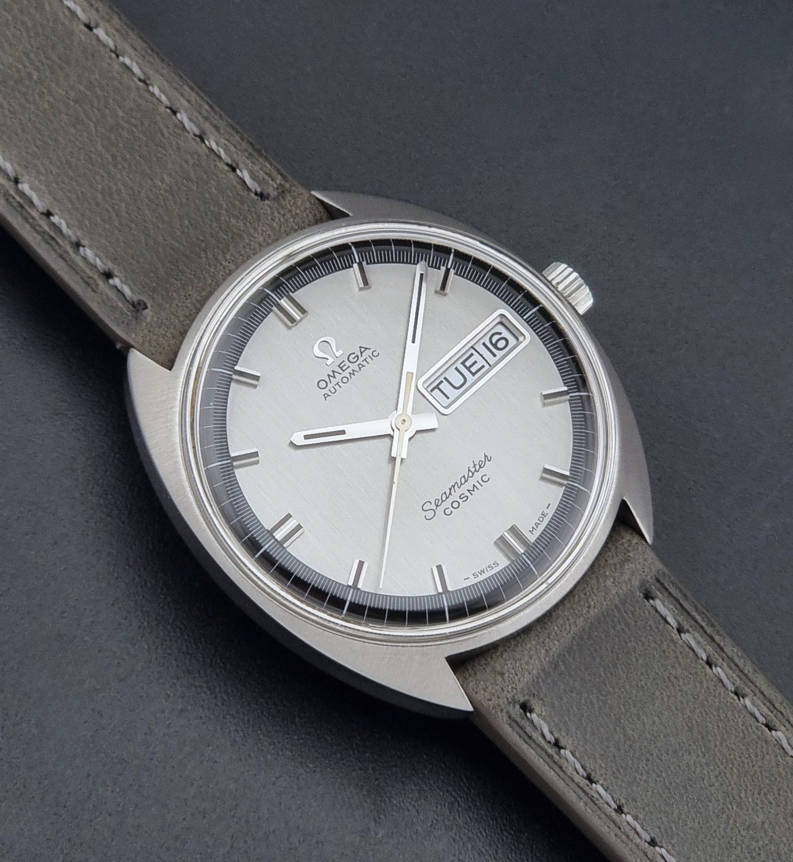 Vintage Omega 166.036 Seamaster Cosmic watch with a silver-toned case, a light gray dial with a day-date window, and a gray leather strap, on a dark background close up dial view