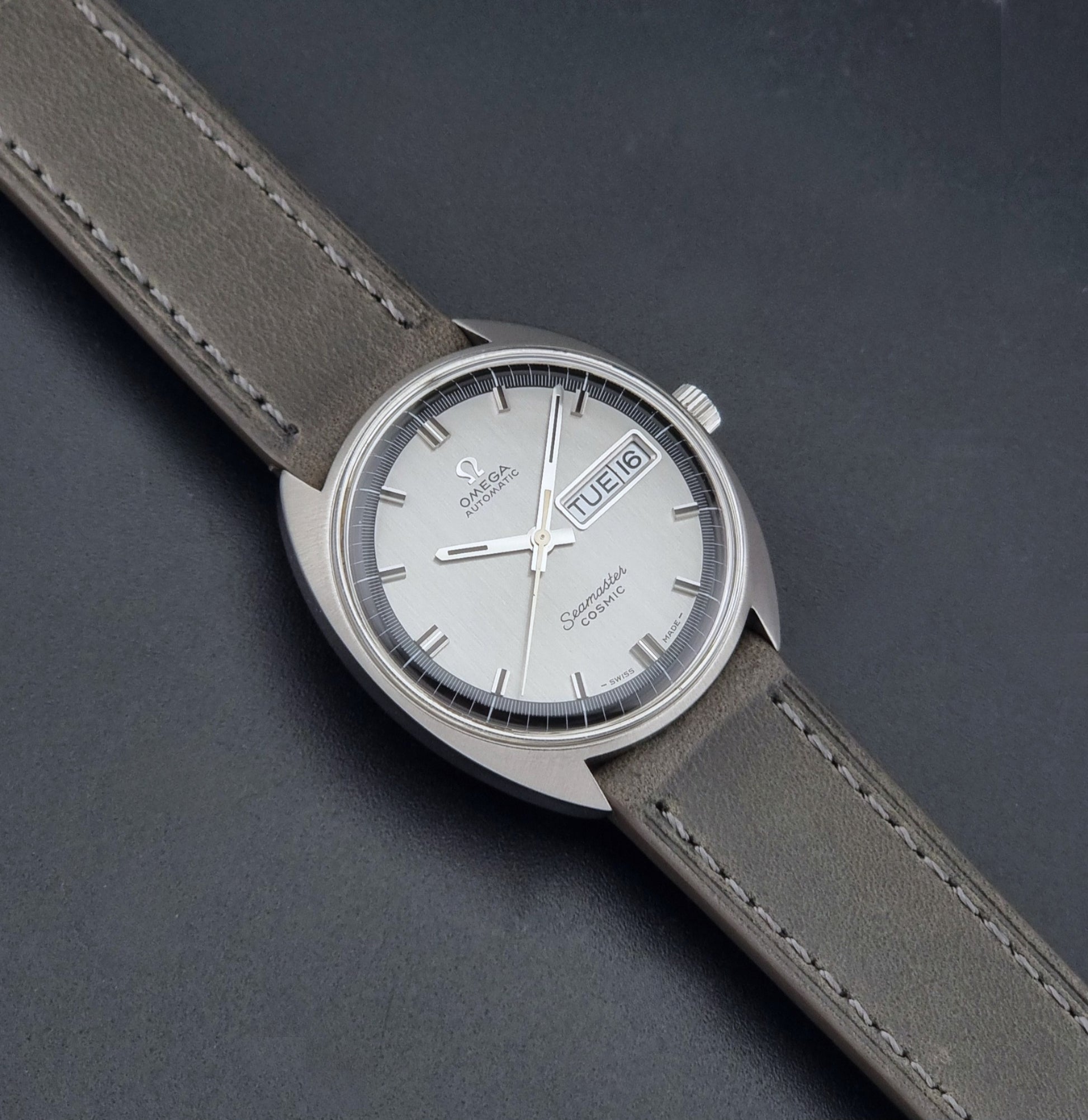 Vintage Omega 166.036 Seamaster Cosmic watch with a silver-toned case, a light gray dial with a day-date window, and a gray leather strap, on a dark background dial