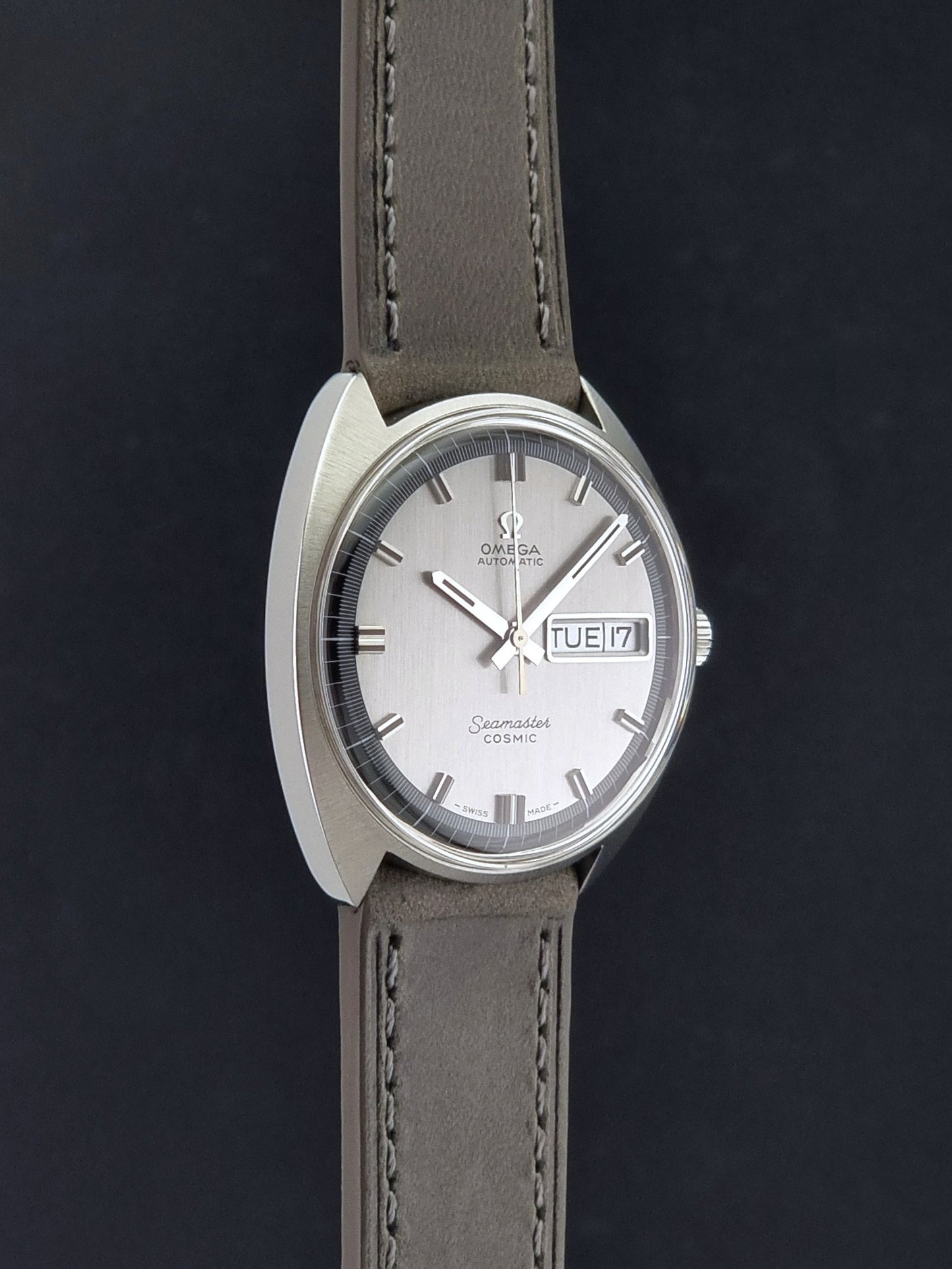 Vintage Omega 166.036 Seamaster Cosmic watch with a silver-toned case, a light gray dial with a day-date window, and a gray leather strap, on a dark background side profile view