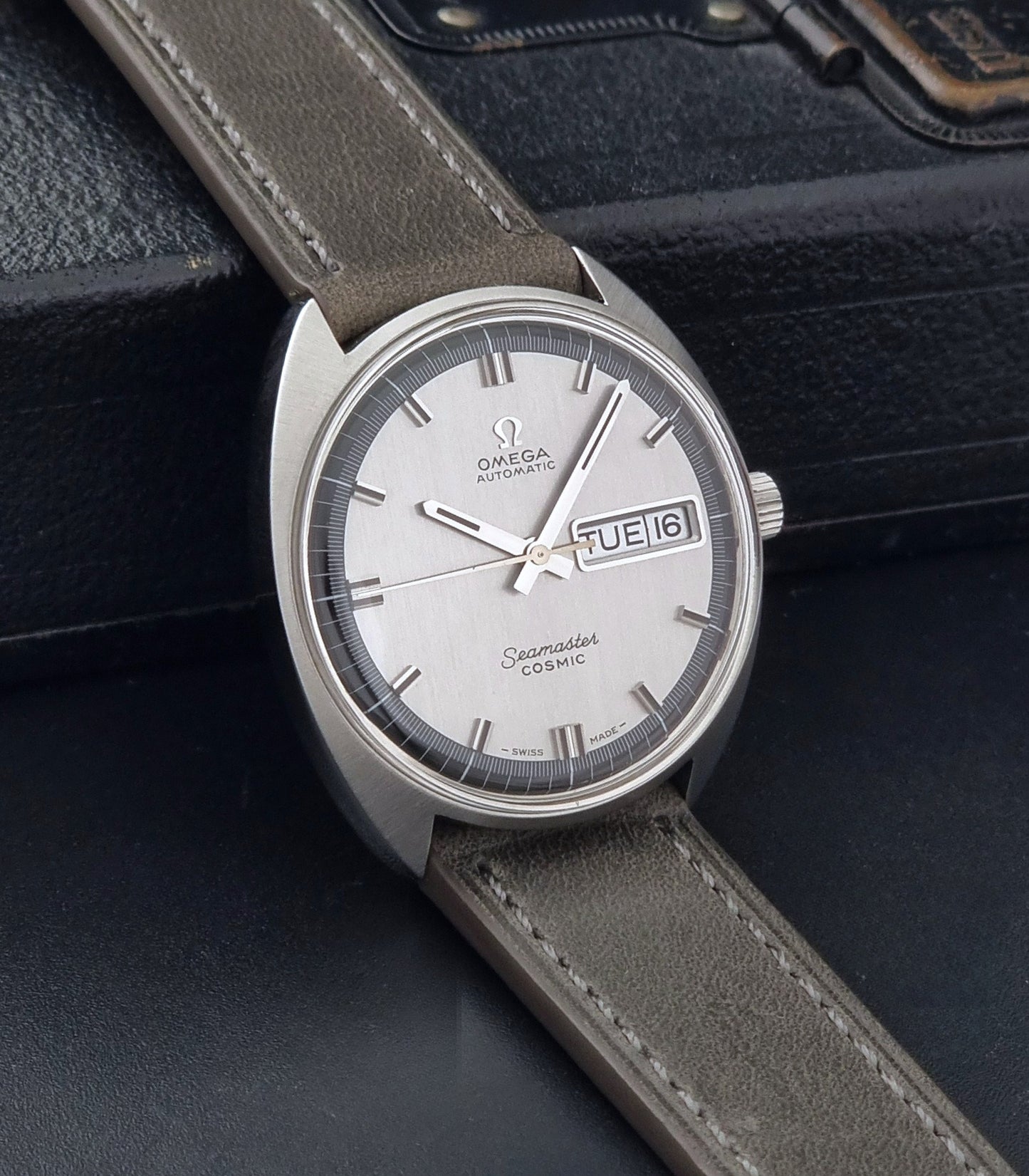Vintage Omega 166.036 Seamaster Cosmic watch with a silver-toned case, a light gray dial with a day-date window, and a gray leather strap, on a dark background side view