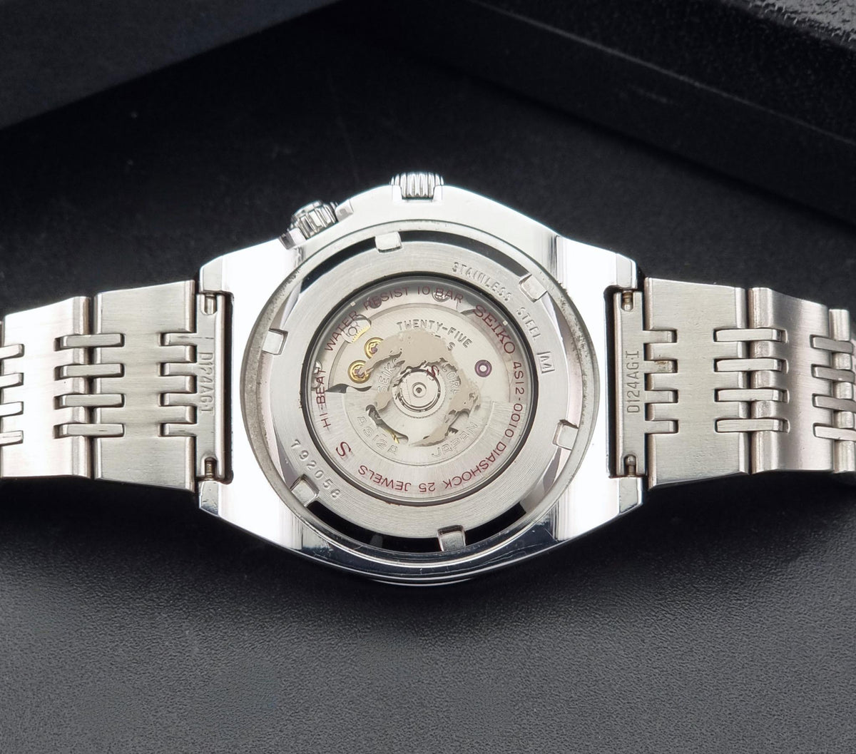 Close-up of the back of a stainless steel Seiko watch Mechasus SCFF007 GMT 4S12-0010 with a transparent case back showing the mechanical movement, on a dark textured surface.