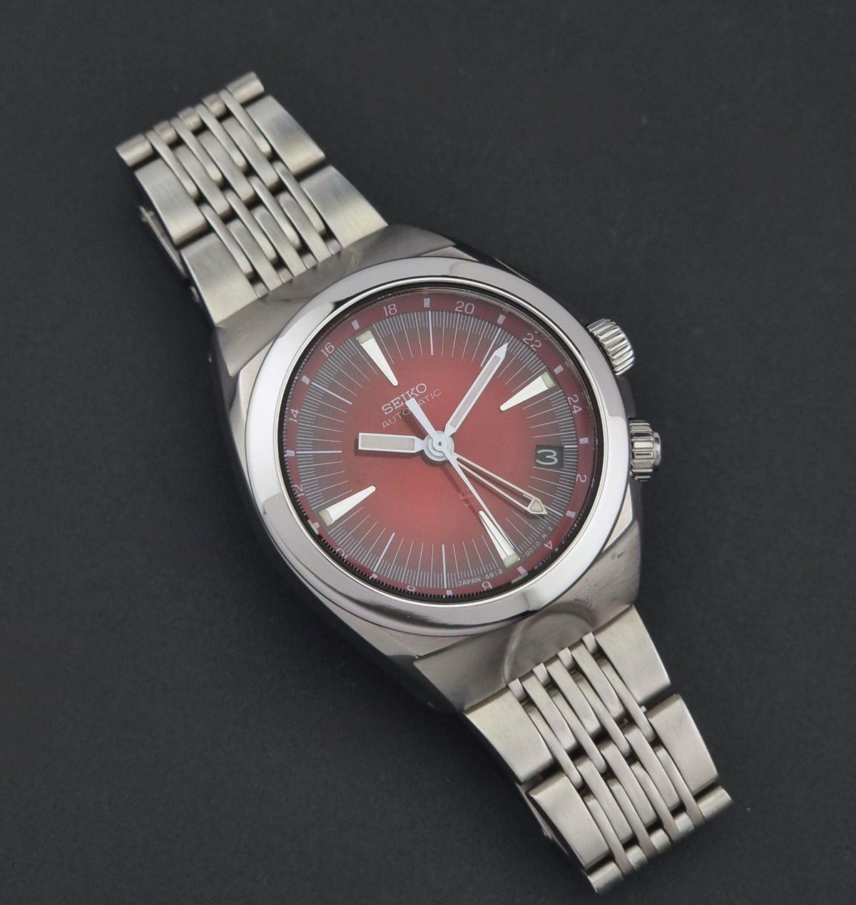 Seiko watch Mechasus SCFF007 GMT 4S12-0010 with a silver-tone metal bracelet and a red sunburst dial, photographed on a dark gray background.