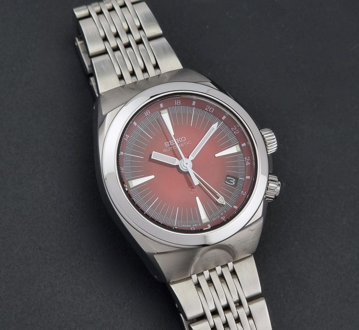 Seiko watch Mechasus SCFF007 GMT 4S12-0010 with a silver-tone metal link bracelet and a red dial, photographed on a dark textured background.