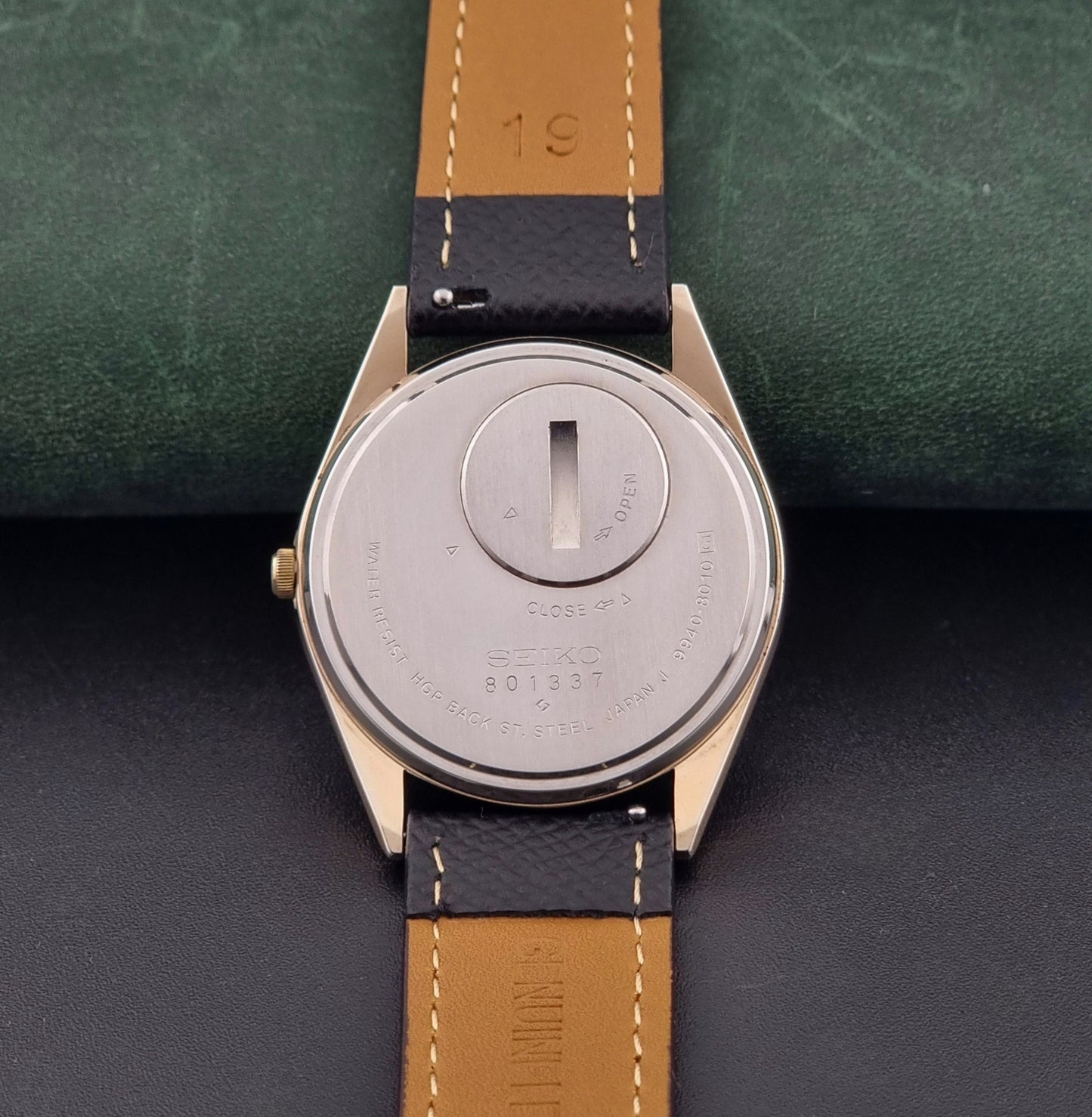 Vintage Seiko Grand Quartz 9940-8010  watch with gold case and black strap on a dark gray background case back view