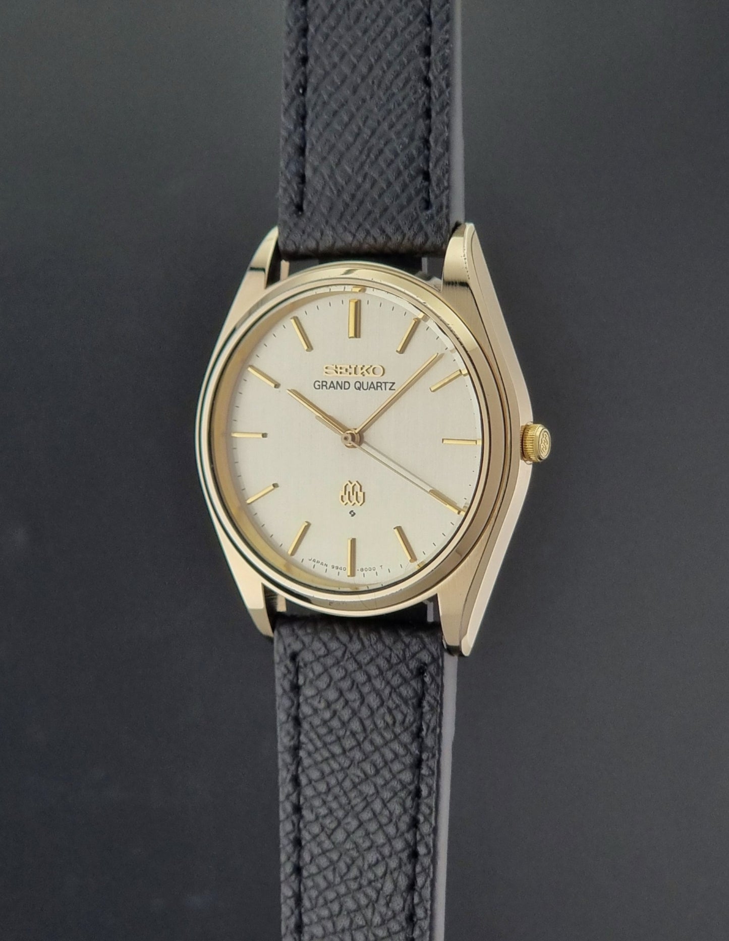 Vintage Seiko Grand Quartz 9940-8010  watch with gold case and black strap on a dark gray background crown profile view