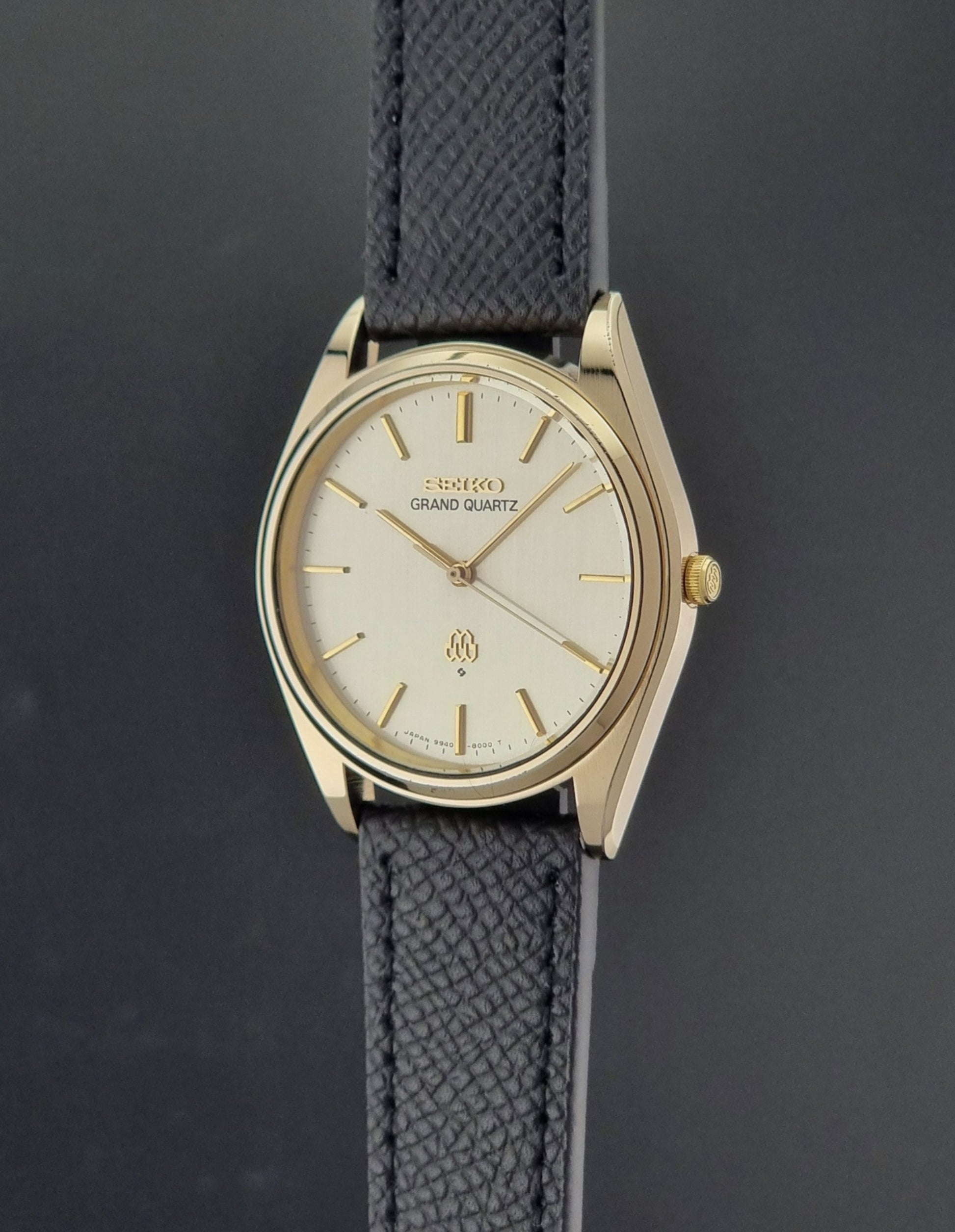 Vintage Seiko Grand Quartz 9940-8010  watch with gold case and black strap on a dark gray background crown profile view