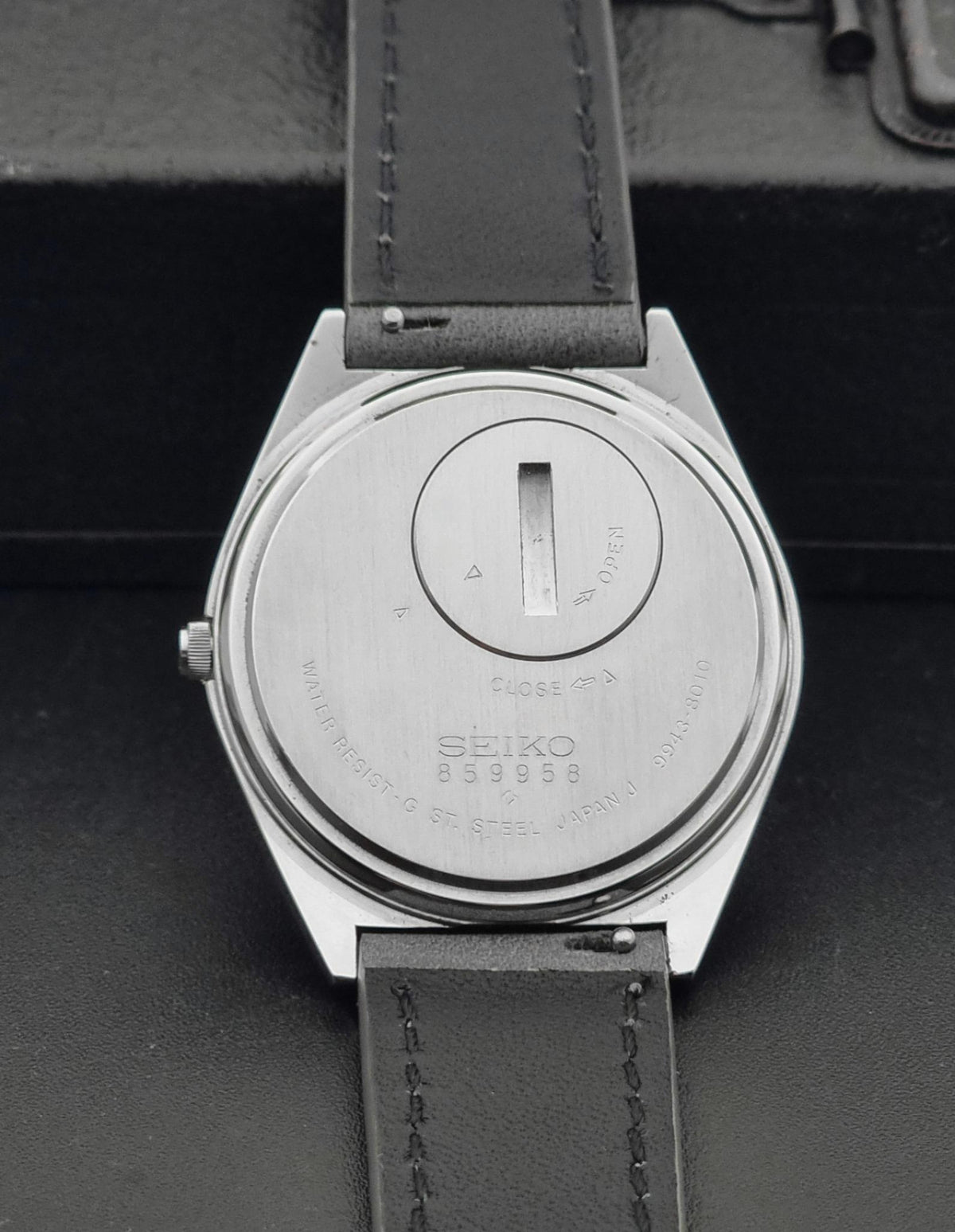 Close-up of the back of a Vintage Seiko Grand Quartz 9943-8010 Wristwatch with a silver-toned metal case and a dark leather strap, resting on a dark textured surface.