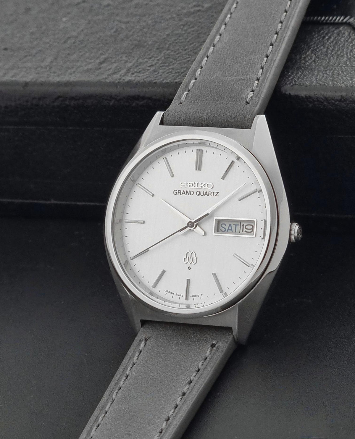 A Seiko Grand Quartz wristwatch with a silver-toned metal case, white dial, and a dark gray leather strap, shown on a dark textured background.