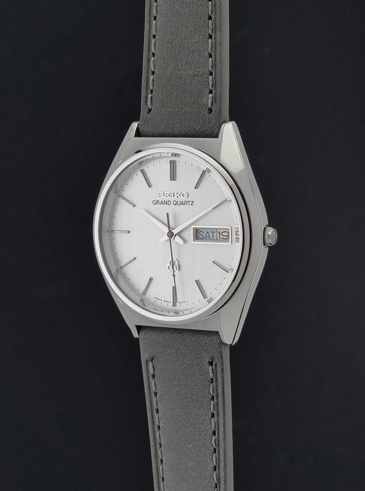 Vintage Seiko Grand Quartz 9943-8010 Wristwatch with a silver-tone case, white dial, and gray leather strap, shown against a dark background.