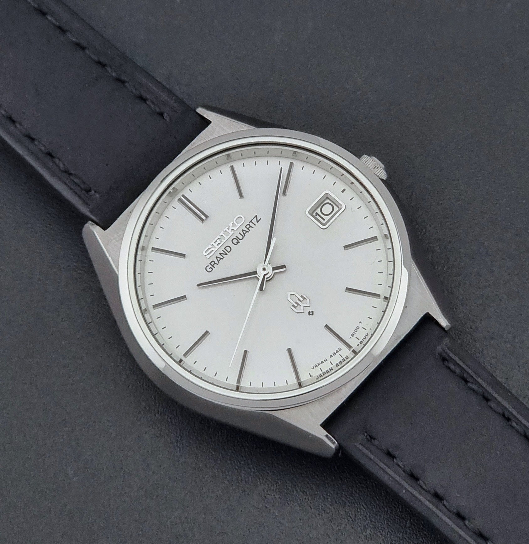 Vintage Seiko Grand Quartz watch 4842-8110 with a silver case and black leather strap on a dark gray background close up dial view