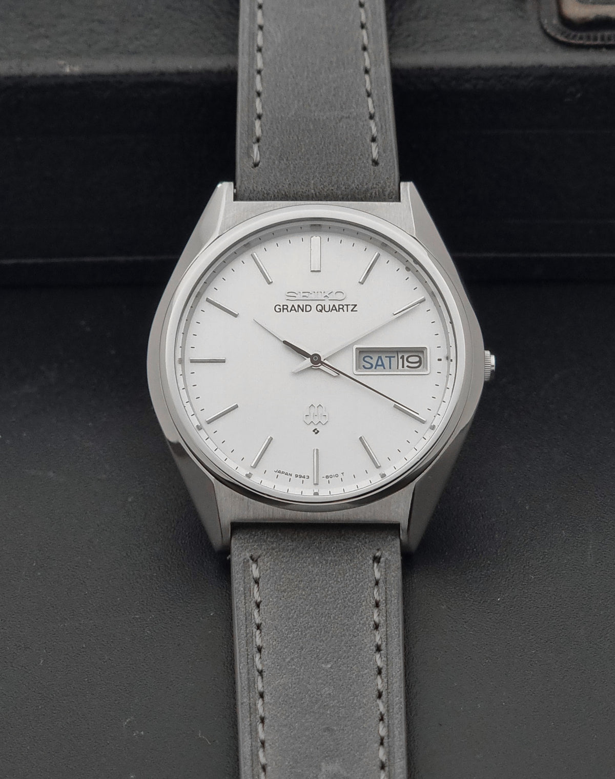 A Seiko Grand Quartz wristwatch with a silver-toned metal case, white dial, and a gray leather strap, displayed on a dark textured surface.