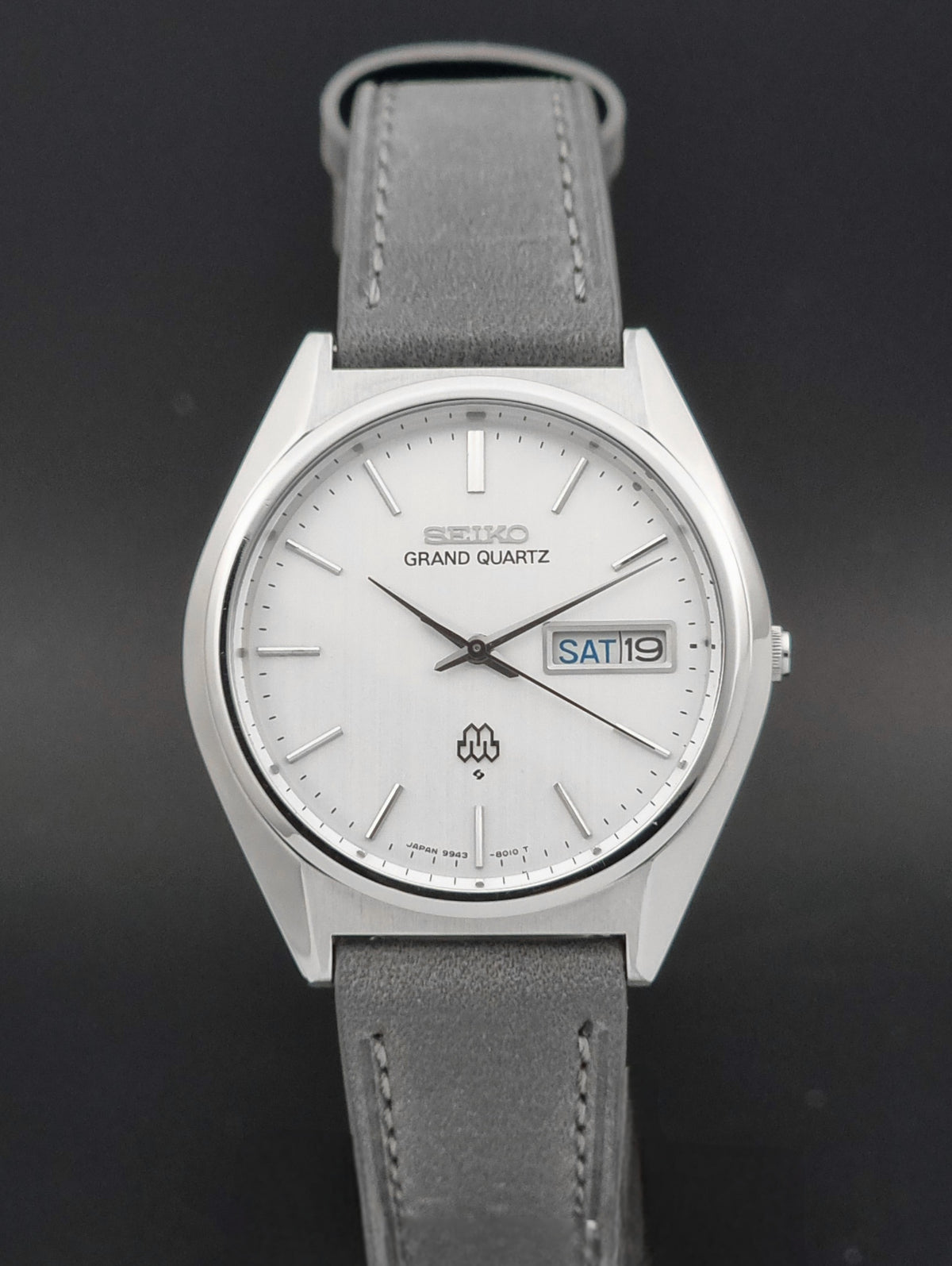 A Seiko Grand Quartz wristwatch with a silver-toned stainless steel case, a white dial, and a gray leather strap, shown against a dark gray background.