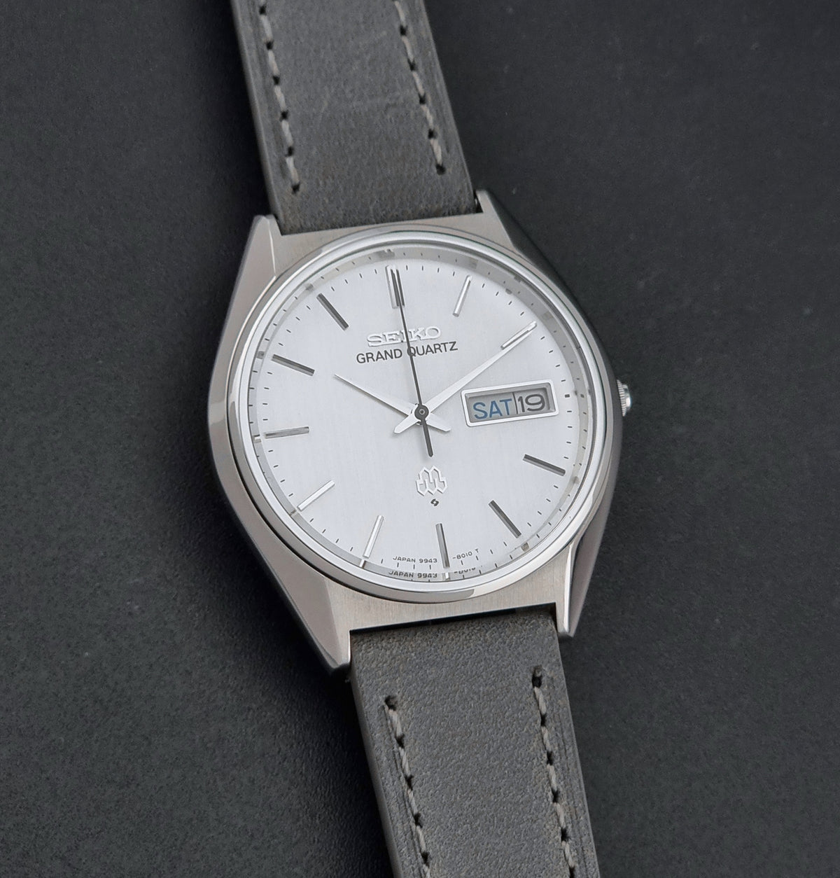 Vintage Seiko Grand Quartz 9943-8010 Wristwatch with a white dial, silver-tone case, and a dark gray leather strap, shown on a dark gray textured background.