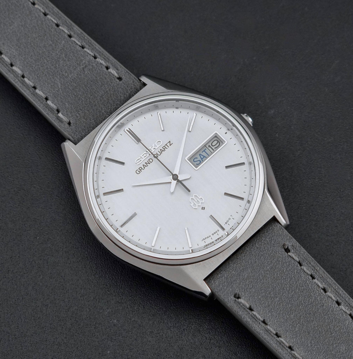 A Seiko Grand Quartz wristwatch with a silver-toned stainless steel case, a white dial with a day-date window, and a dark gray leather strap, shown on a dark textured background.