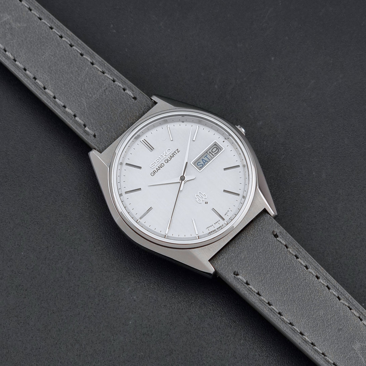 A Seiko Grand Quartz wristwatch with a silver-toned metal case, white dial, and gray leather strap, shown diagonally on a dark textured background.