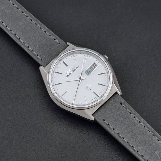 A Seiko Grand Quartz wristwatch with a silver-toned metal case, white dial, and gray leather strap, shown diagonally on a dark textured background.