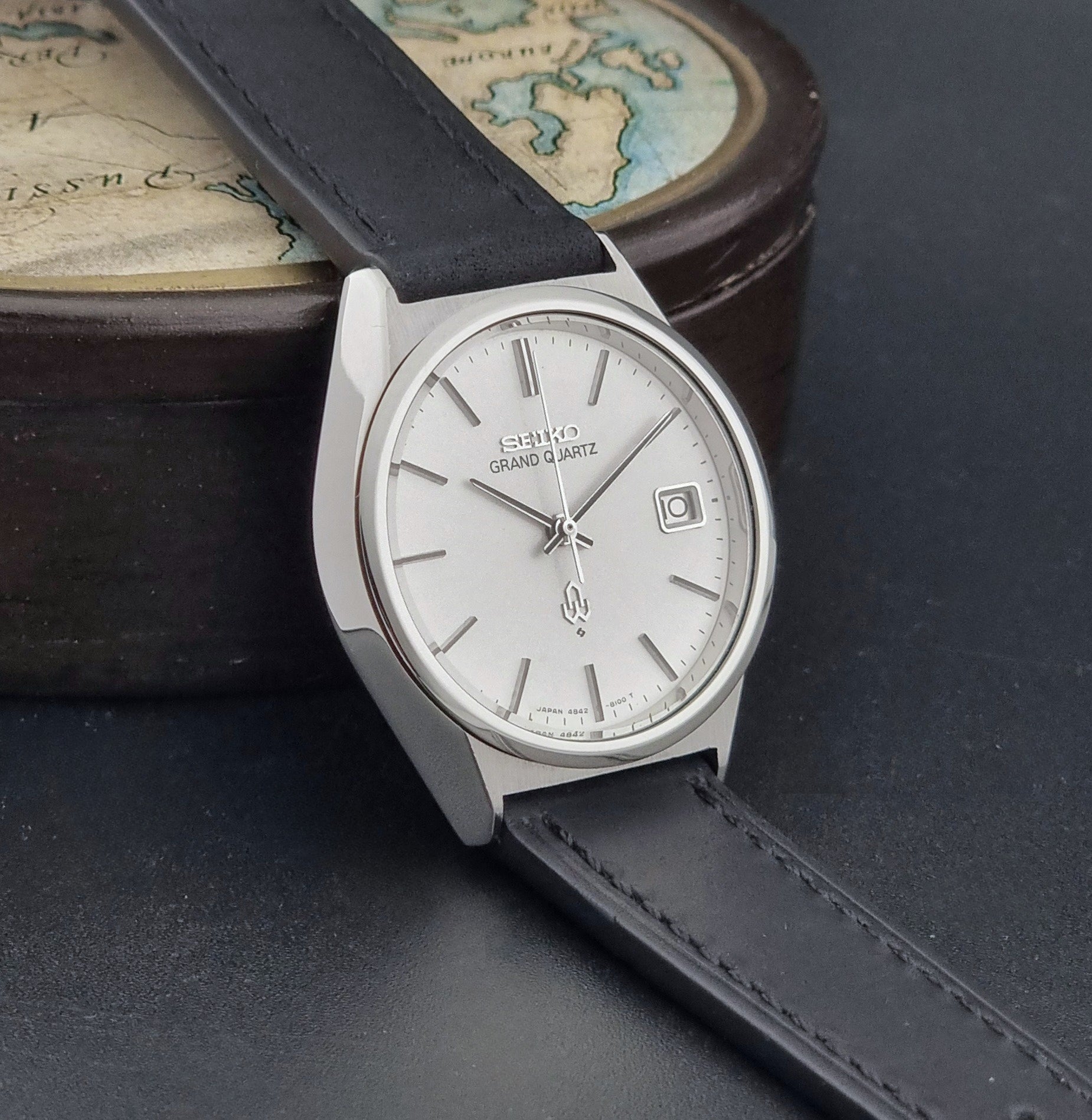 Vintage Seiko Grand Quartz watch 4842-8110 with a silver case and black leather strap on a dark gray background side view